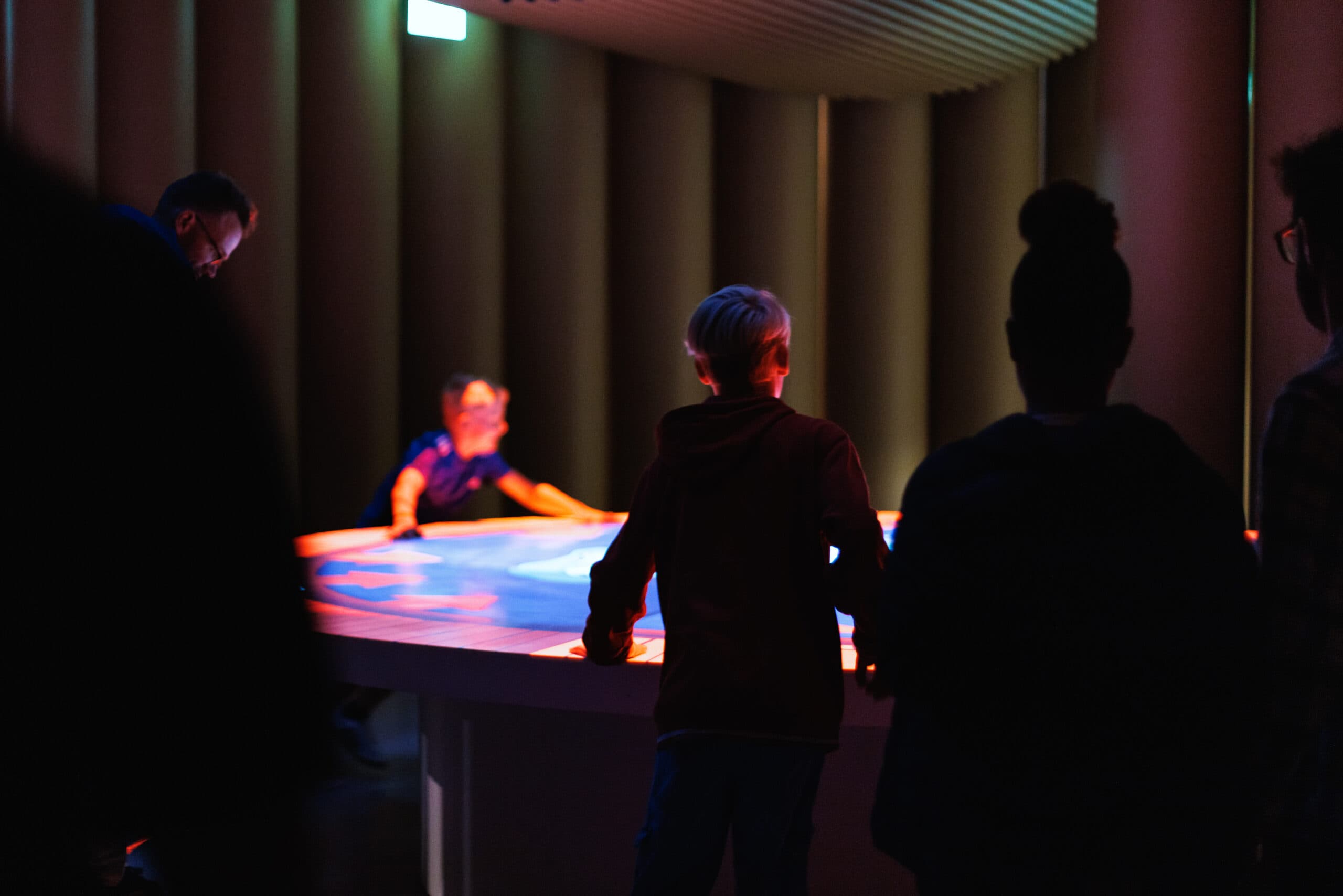 Plusieurs visiteurs sont dans une pièce sombre, autour d'une table lumineuse sur laquelle est projeté un jeu interactif.