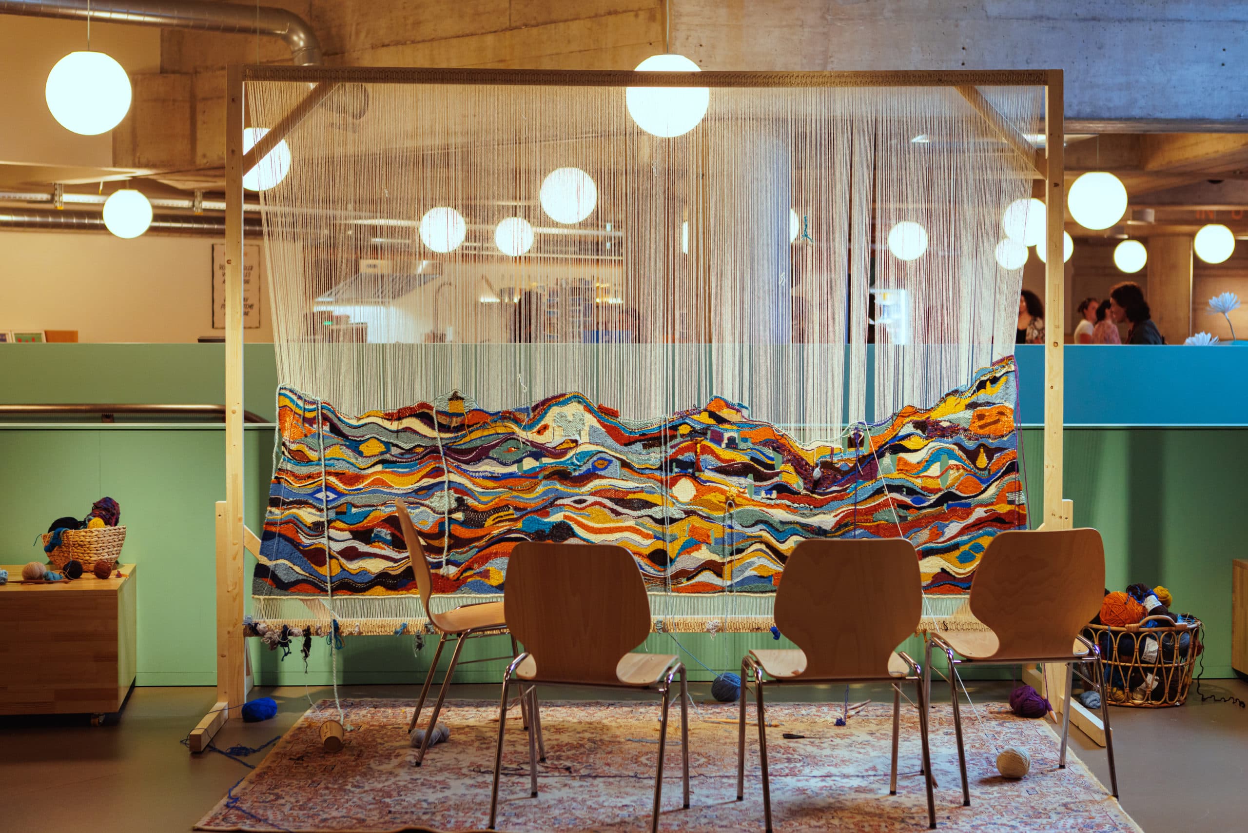 A wooden loom set up indoors, with a large, colourful tapestry featuring wavy patterns currently being woven, balls of wool on the floor and three chairs arranged in front of it.
