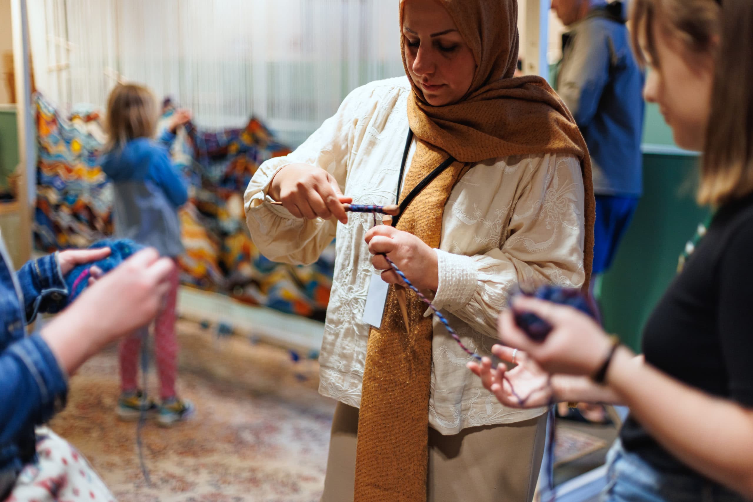 A close-up of the hands of three women spinning wool. In the background, a little girl is weaving a colourful tapestry on a large vertical loom.
