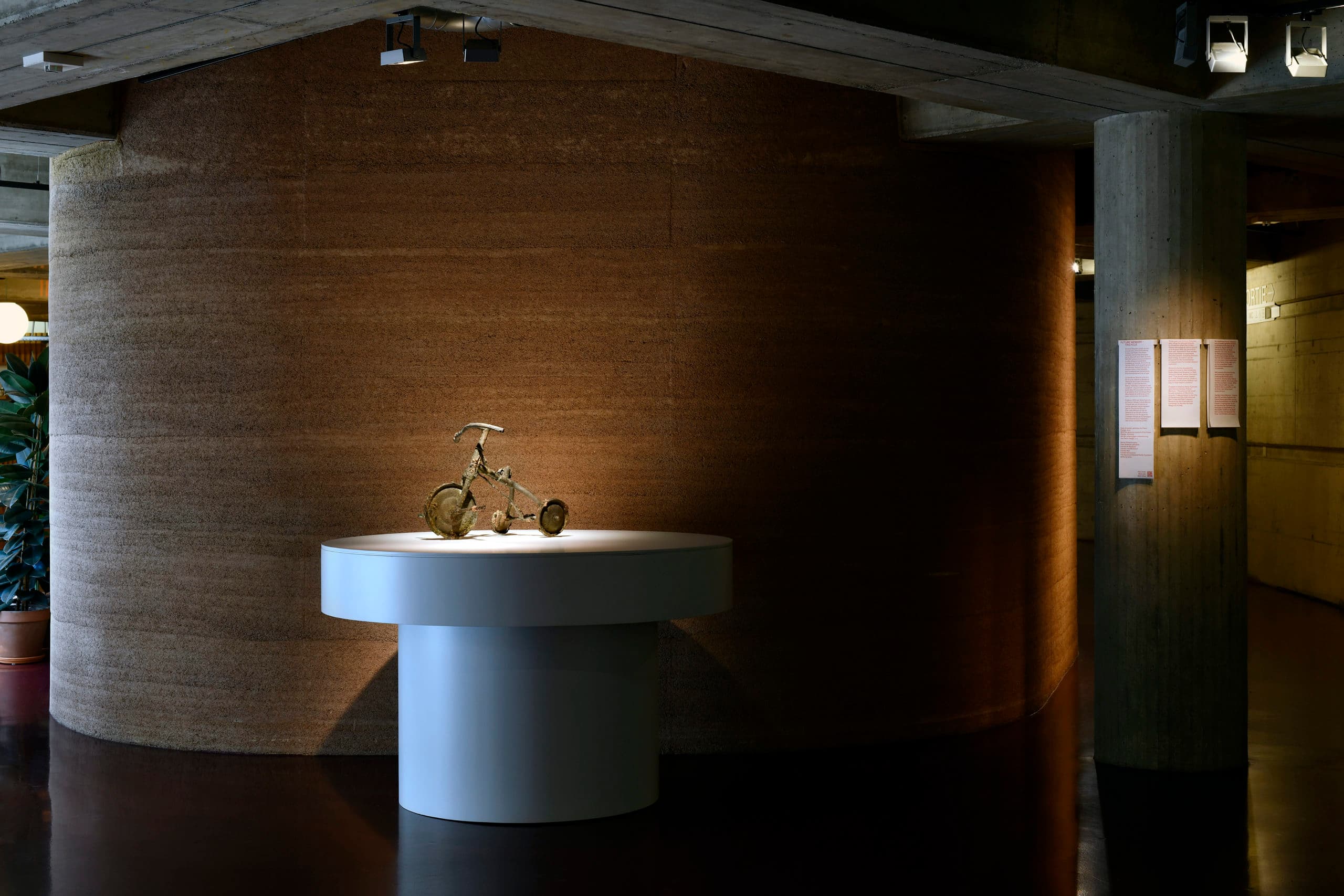 Sculpture d'un tricycle en métal posé sur un socle blanc avec un éclairage direct, devant un mur cylindrique marron et texturé.