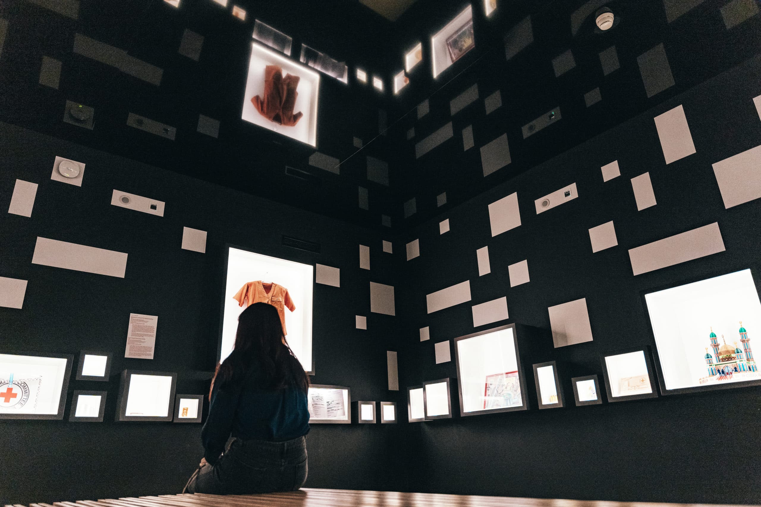 Eine Person, die mit dem Rücken zur Kamera sitzt, betrachtet einen Ausstellungsraum mit dunklen Wänden, an denen zahlreiche Bilderrahmen und Leuchtbildschirme unterschiedlicher Größe angebracht sind, auf denen Objekte und Illustrationen zu sehen sind.