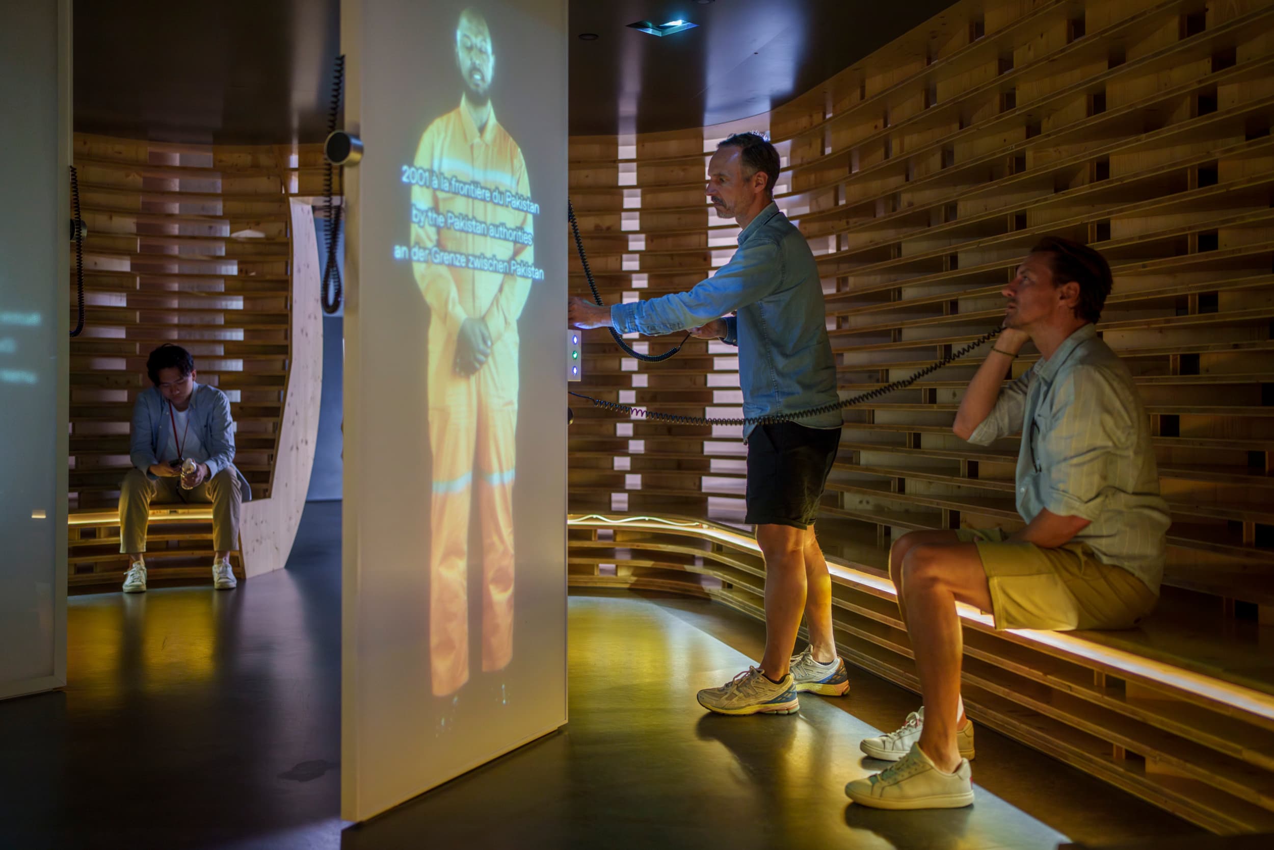 Deux visiteurs écoutent avec des casques le témoignage projeté sur un écran d'un homme en tenue de travail orange.