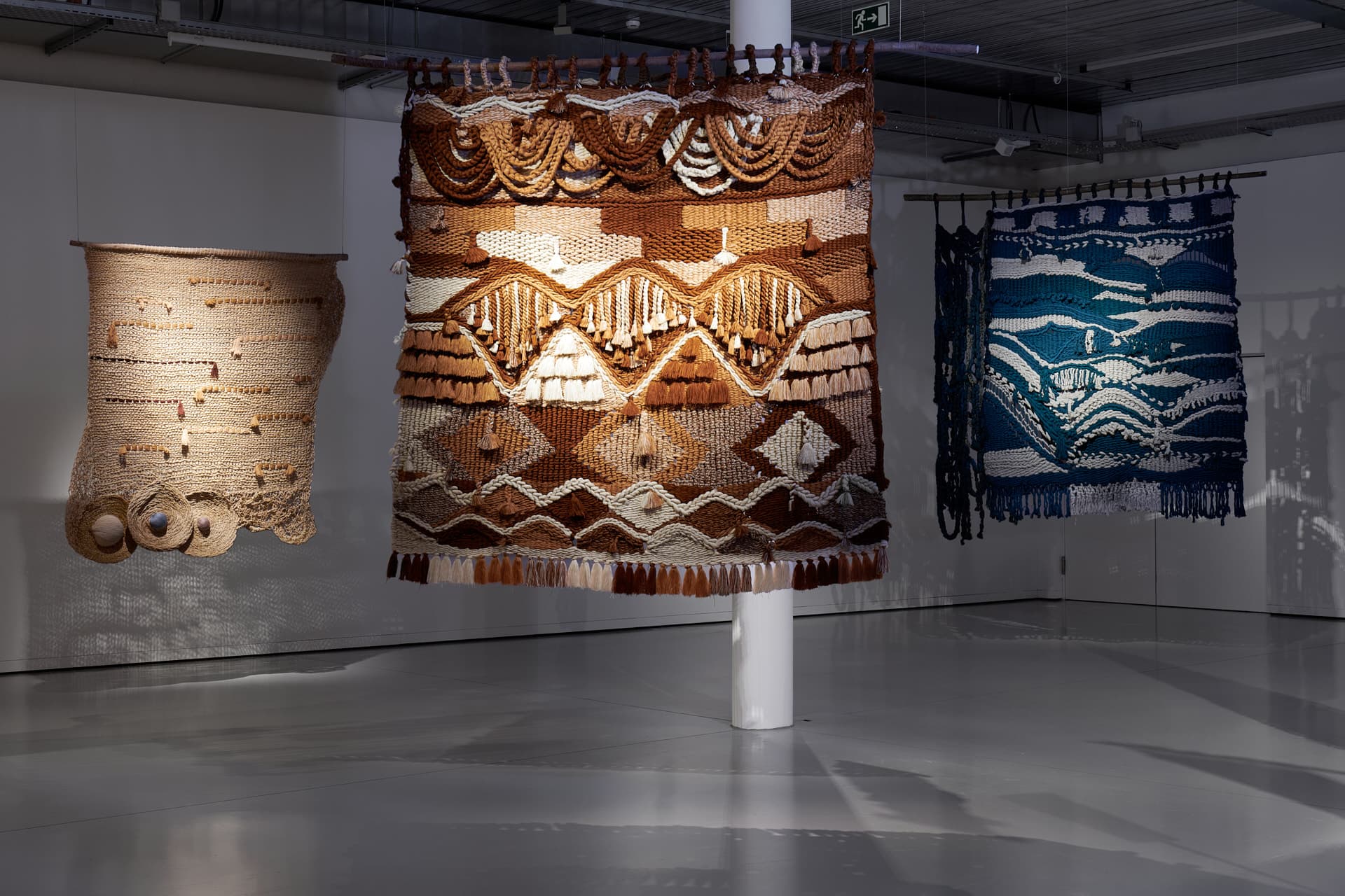 Grande tapisserie suspendue aux tons bruns, beiges et crème, composée de motifs géométriques et de franges, exposée aux côtés d’autres œuvres similaires.