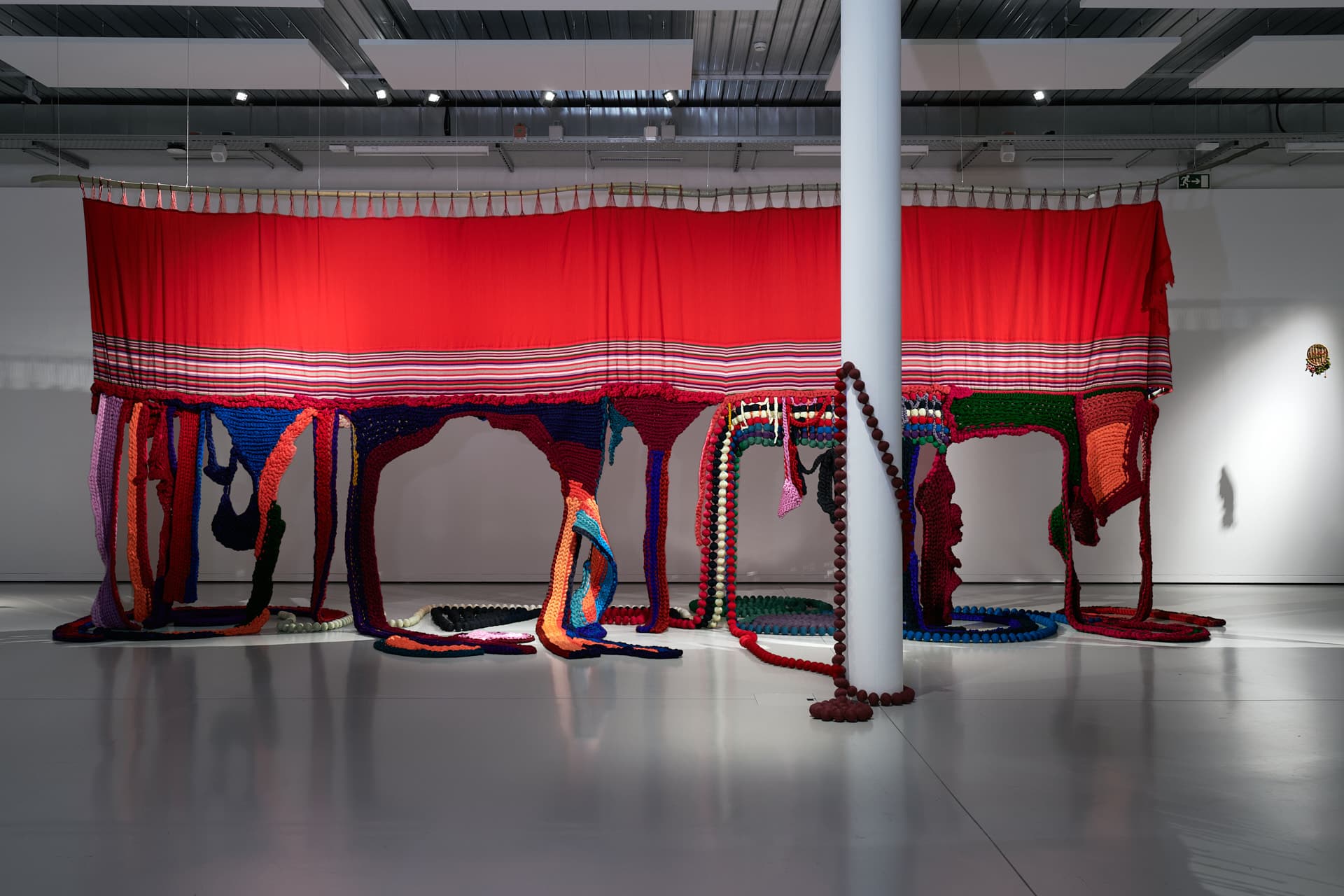 Prise de vue de l’œuvre "Germinar desde las profundas raices". Une grande installation textile rouge est suspendue et des franges et des éléments de tissage colorés tombent jusqu’au sol dans un espace d’exposition.