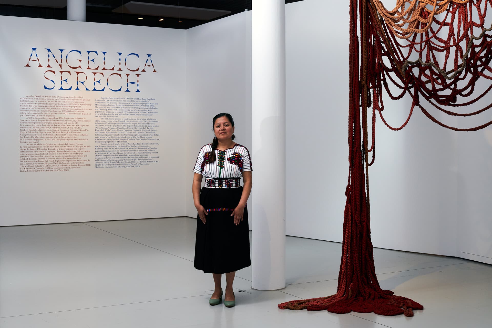 Portrait of the artist Angélica Serech standing in an exhibition space next to a large red textile installation and a wall displaying the title “Angélica Serech” and an explanatory text.