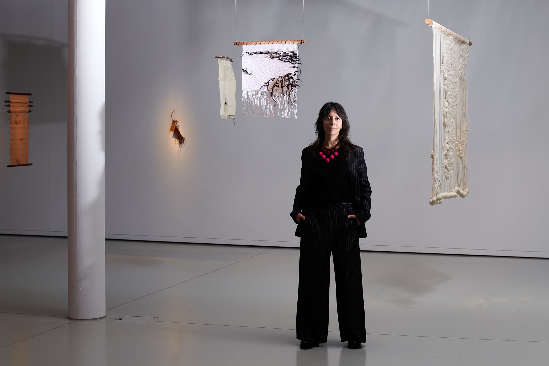 Portrait of the curator Elisa Rusca. A woman dressed in black stands in a minimalist exhibition space, surrounded by suspended textile pieces in light colours.