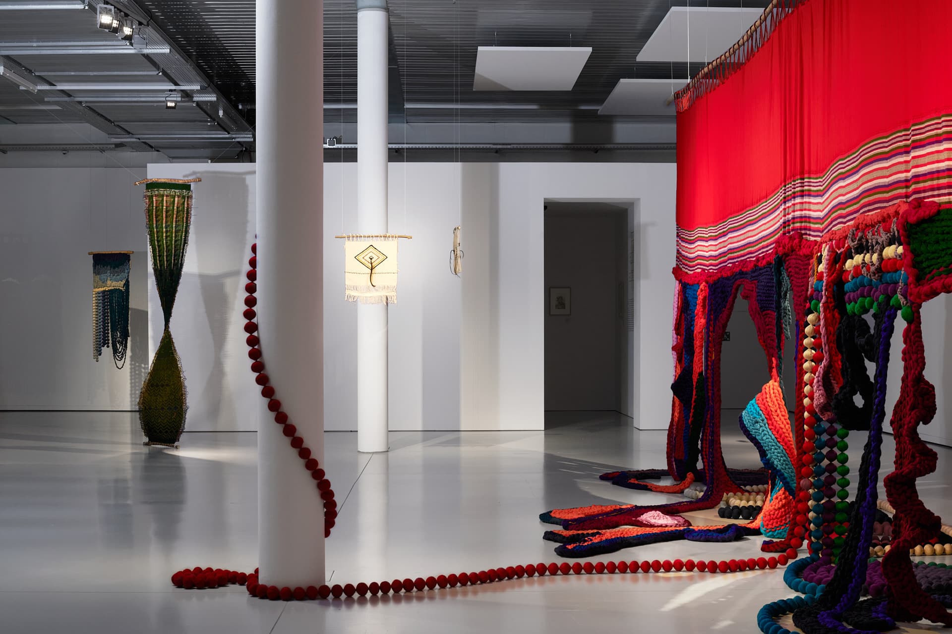 A bright exhibition space with white columns, where a red and multicoloured textile installation spreads across the floor and along a wall, accompanied by other suspended pieces in the background.