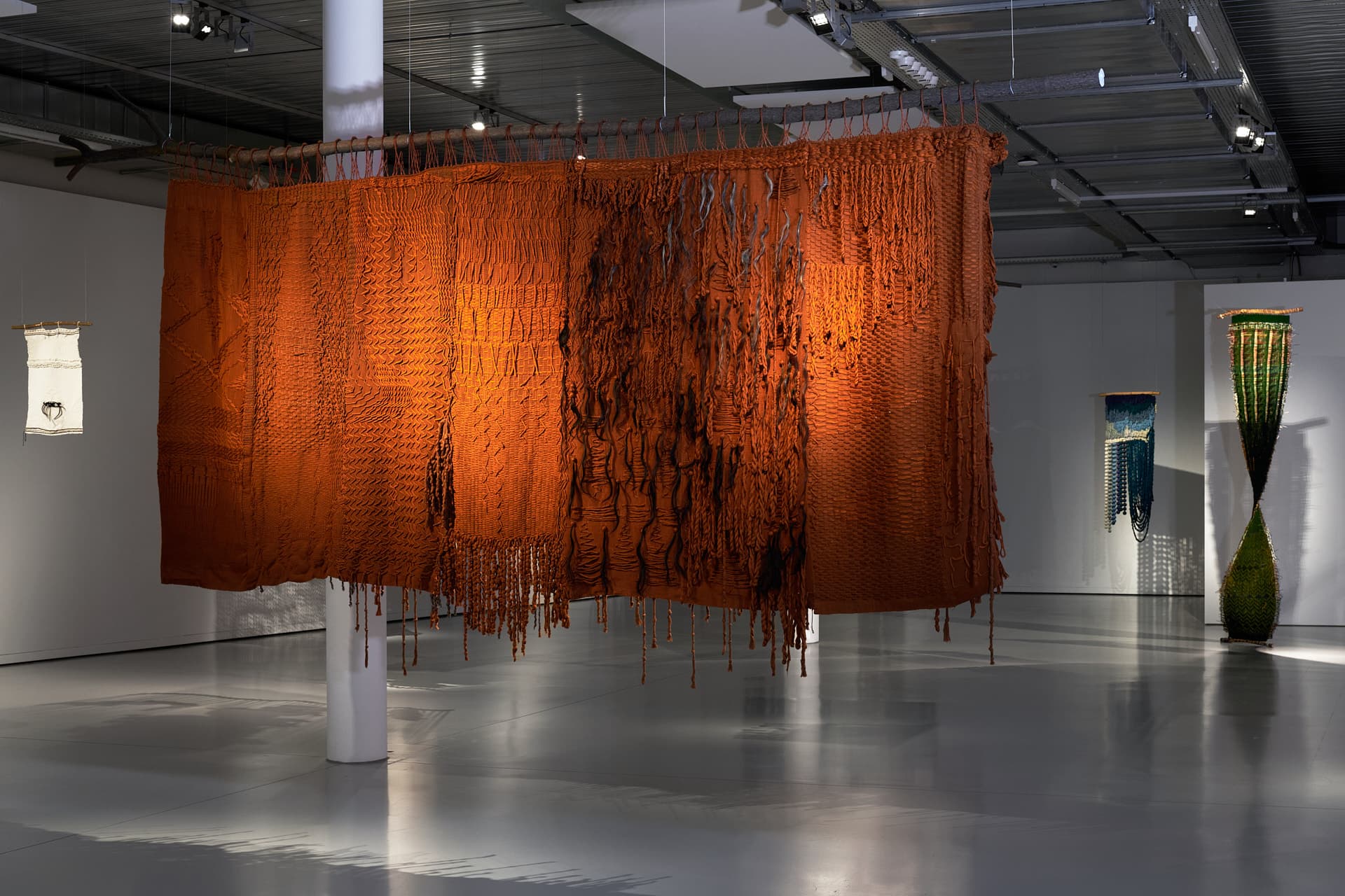 Prise de vue de l’œuvre "Mi historia en nudos, al dorso de mi Güipil" Une large installation textile brun-orangé suspendue au centre d’une galerie, composée de nœuds, de franges tombantes, et de mèches e cheveux noirs et gris éclairée par des spots.