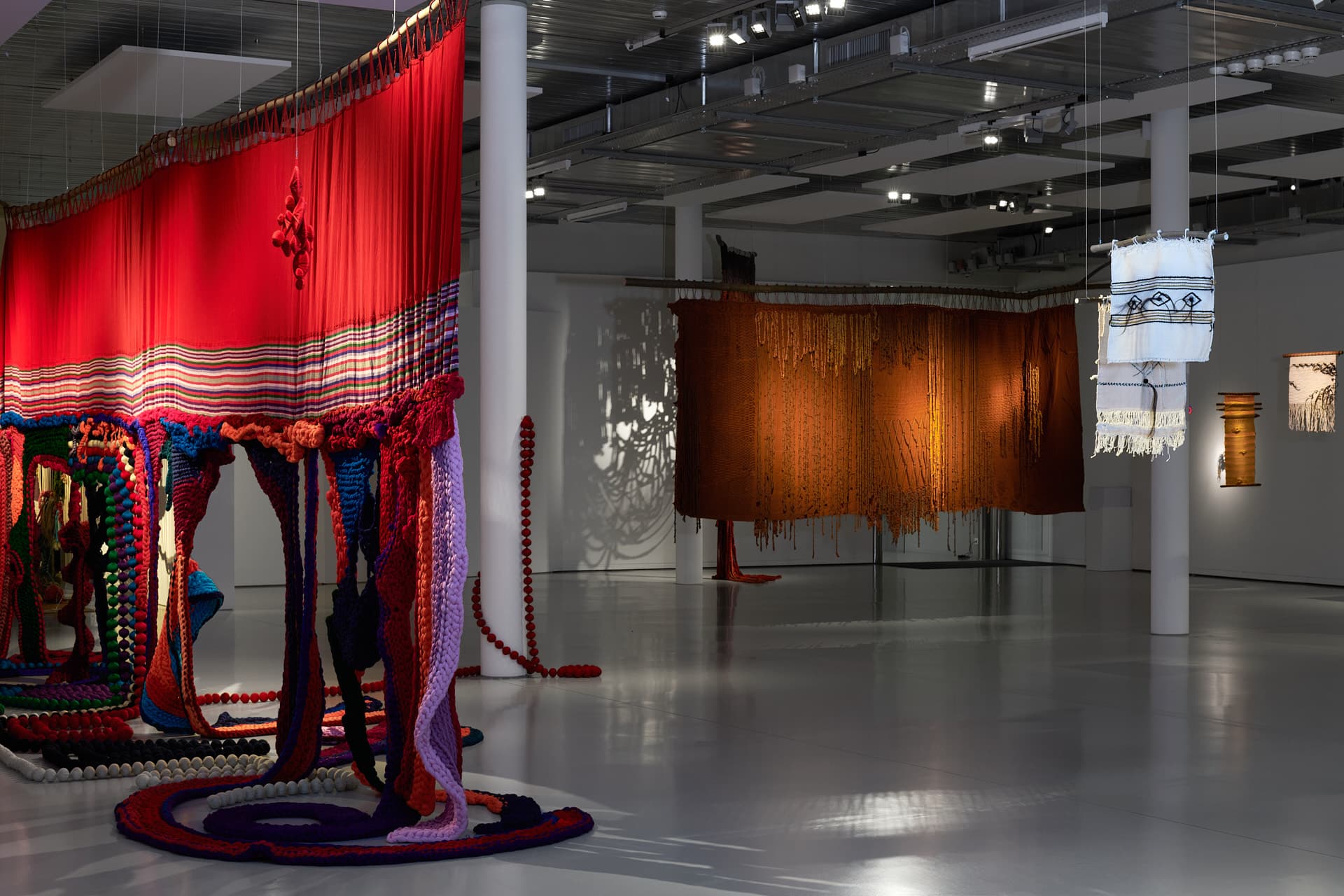 Des tapisseries rouge, brun-orangé et blanche sont suspendues depuis le plafond d'une salle d'exposition