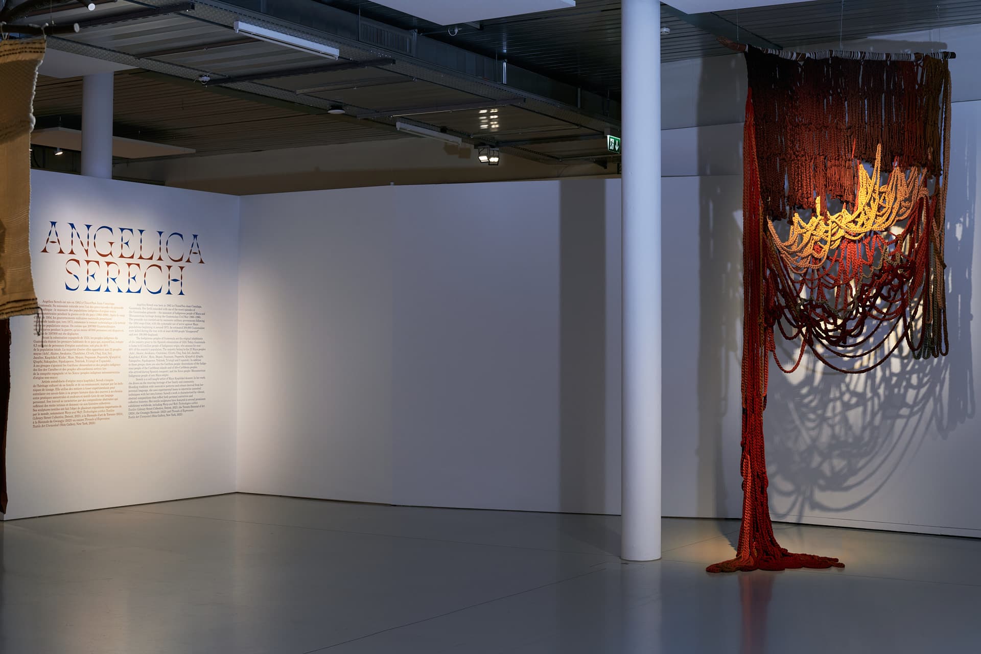 Prise de vue de l’œuvre "Sororidad", une tapisserie de de couleur ocre et beige avec des tressages ocres qui tombent jusqu'au sol. Sur la gauche, un mur blanc avec une notice de présentation de l'exposition et le nom de l'artiste.