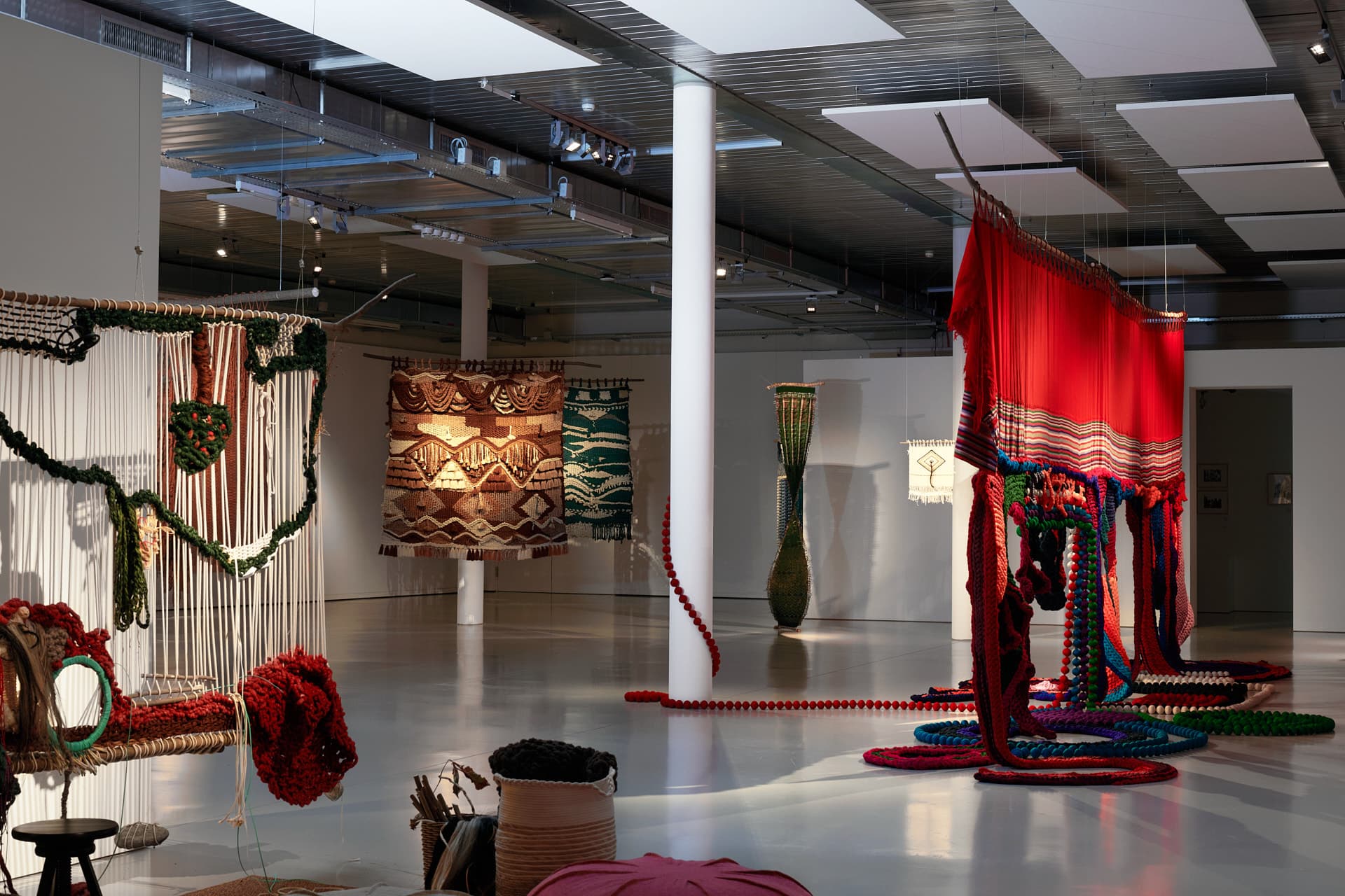 Overview of an exhibition hall with several colourful textile installations suspended from the ceiling, featuring works with geometric patterns and brightly coloured elements such as red and green.