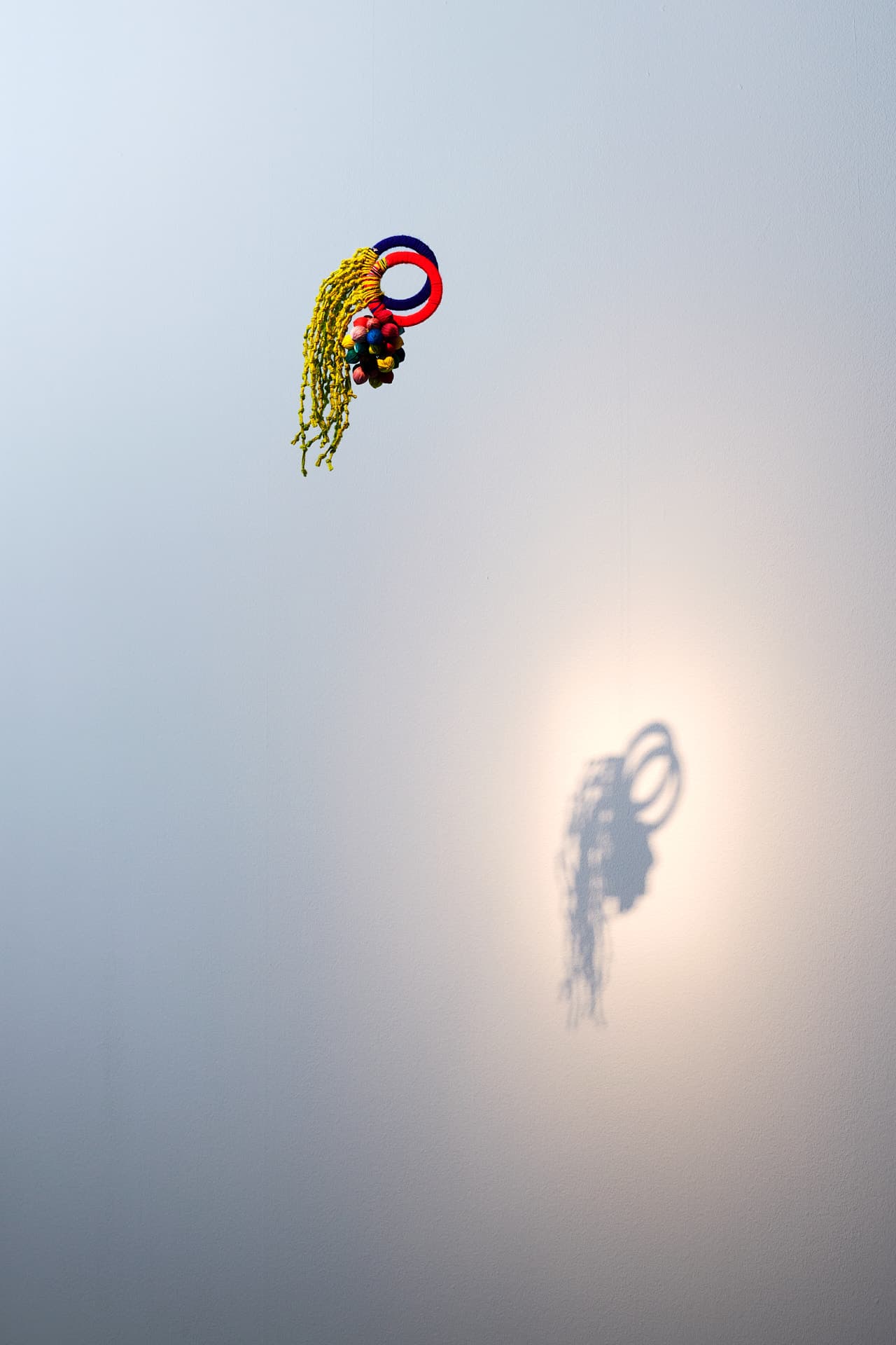 Photograph of “Alegría en Chixot / Comalapa”. A suspended textile installation comprising two circular shapes in red and blue, to which threads forming knots and small balls are attached, casting a shadow onto a light-coloured wall.
