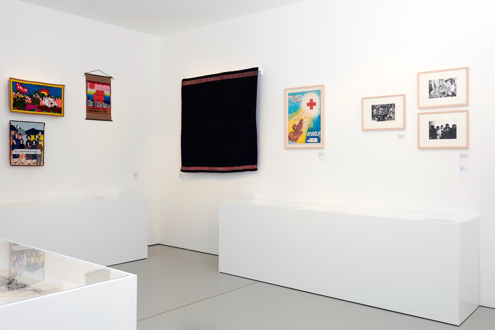 Exhibition hall with white walls displaying a dark tapestry, black-and-white photographs and a poster. On the left-hand wall, three small colourful textile works are hung.