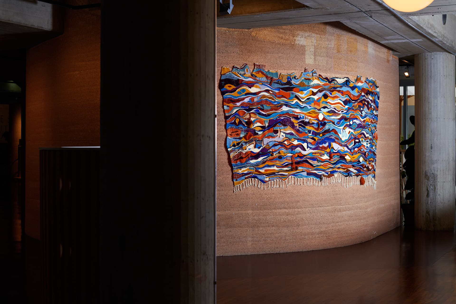 Tapisserie aux motifs ondulés multicolores (bleu, orange, rouge, blanc), suspendue sur un mur incurvé en chanvre texturé.