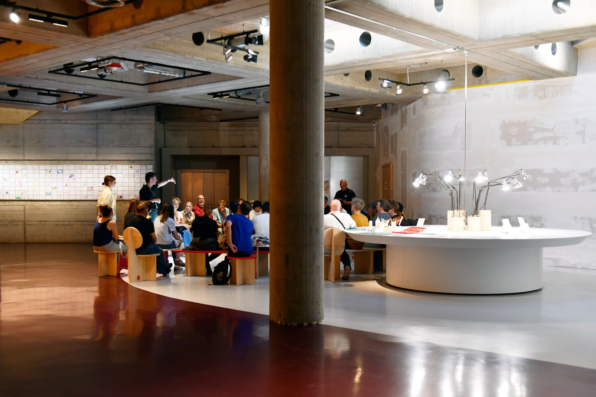 Un groupe de personnes assis en cercle dans un espace lumineux en sous-sol. Derrière eux, une grande table blanche et circulaire avec de nombreuses lampes au centre.