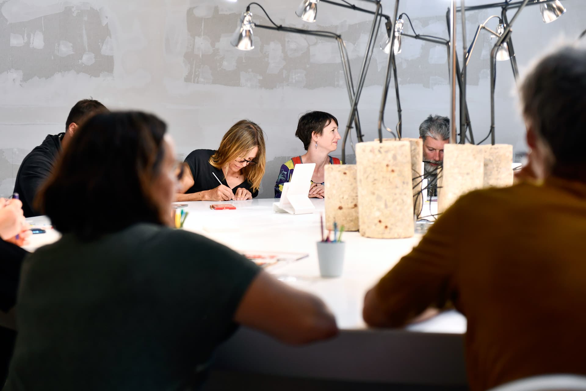 Menschen, die um einen weißen Tisch sitzen und mit Zeichenmaterial zeichnen, während in der Mitte des Tisches Lampen stehen. Im Vordergrund sind zwei Silhouetten von Personen im Gegenlicht zu sehen, die um diesen Tisch sitzen.