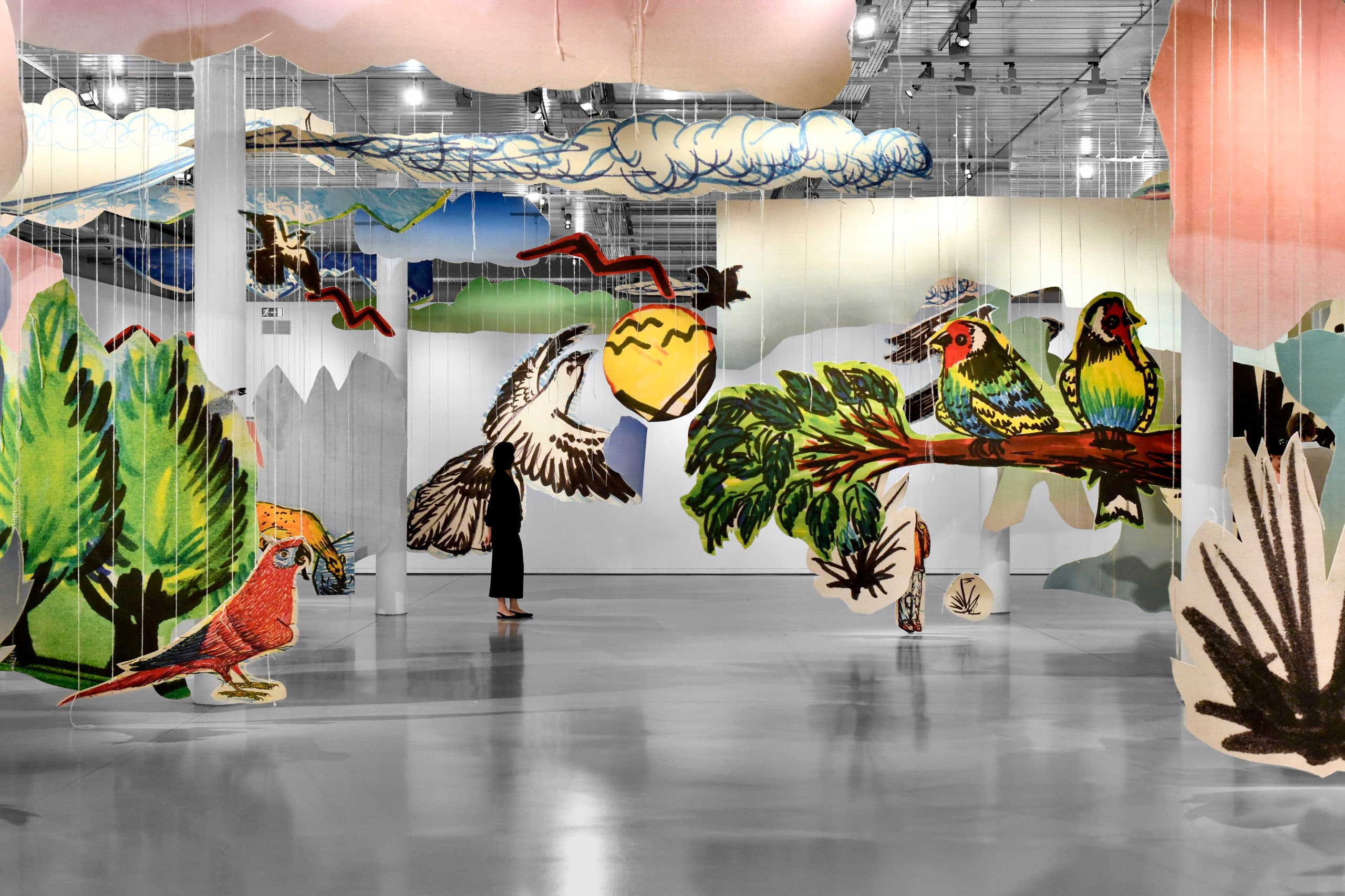 An art installation featuring giant, colourful cut-outs of birds, branches and foliage suspended in mid-air, with visitors walking amongst the cut-outs.