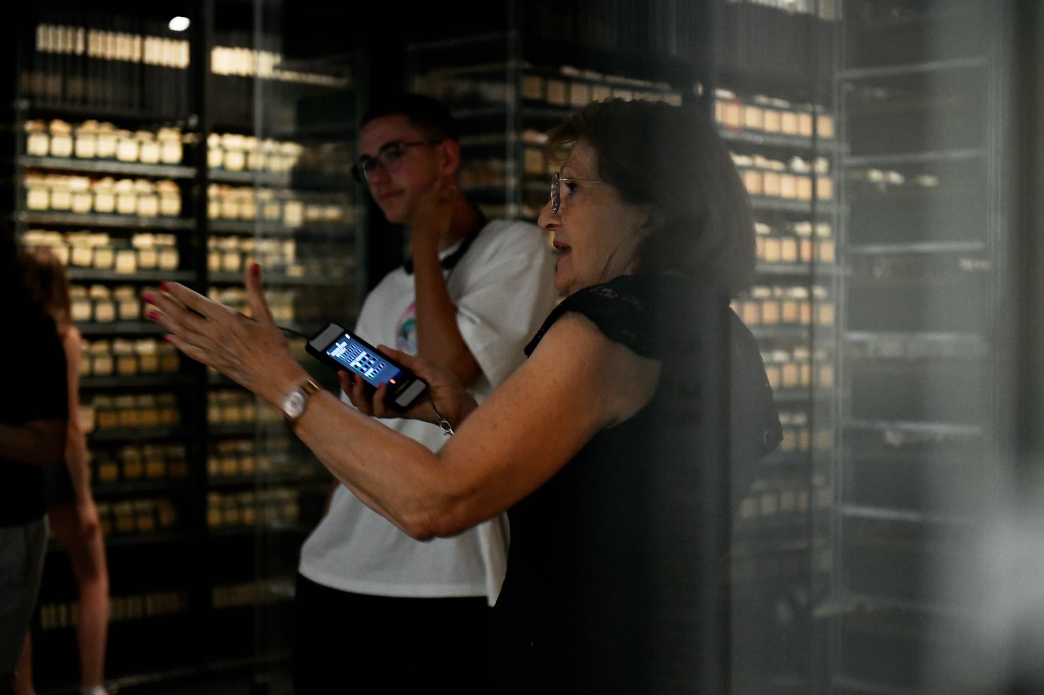 Une femme et un adolescent se tiennent debout dans une salle, autour d'eux des vitrines remplies d'archives. La femme signale quelque chose du bras que l'adolescent regarde.