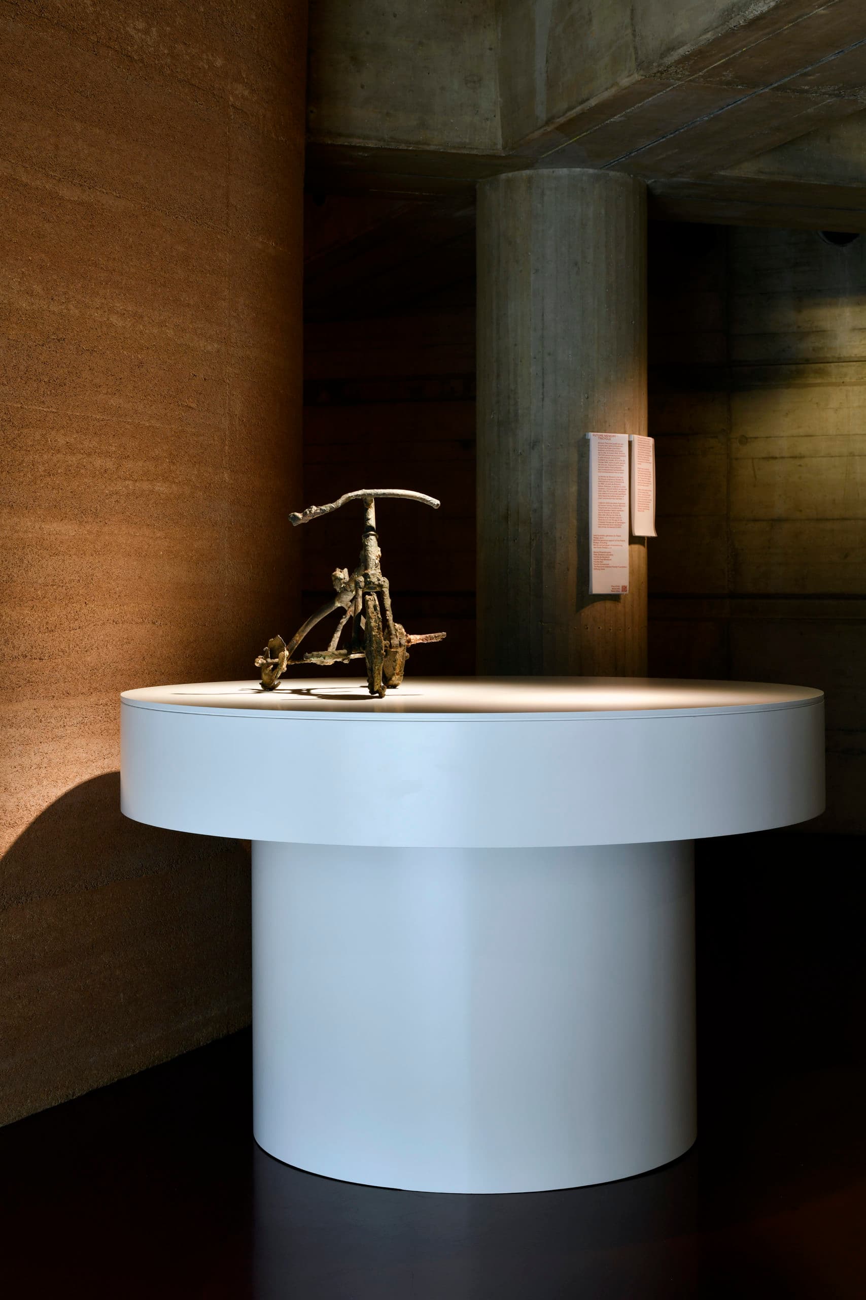 A metal sculpture of a tricycle displayed on a large white circular plinth, illuminated by a spotlight. In the background, a textured brown wall and a concrete column bearing an explanatory sign.