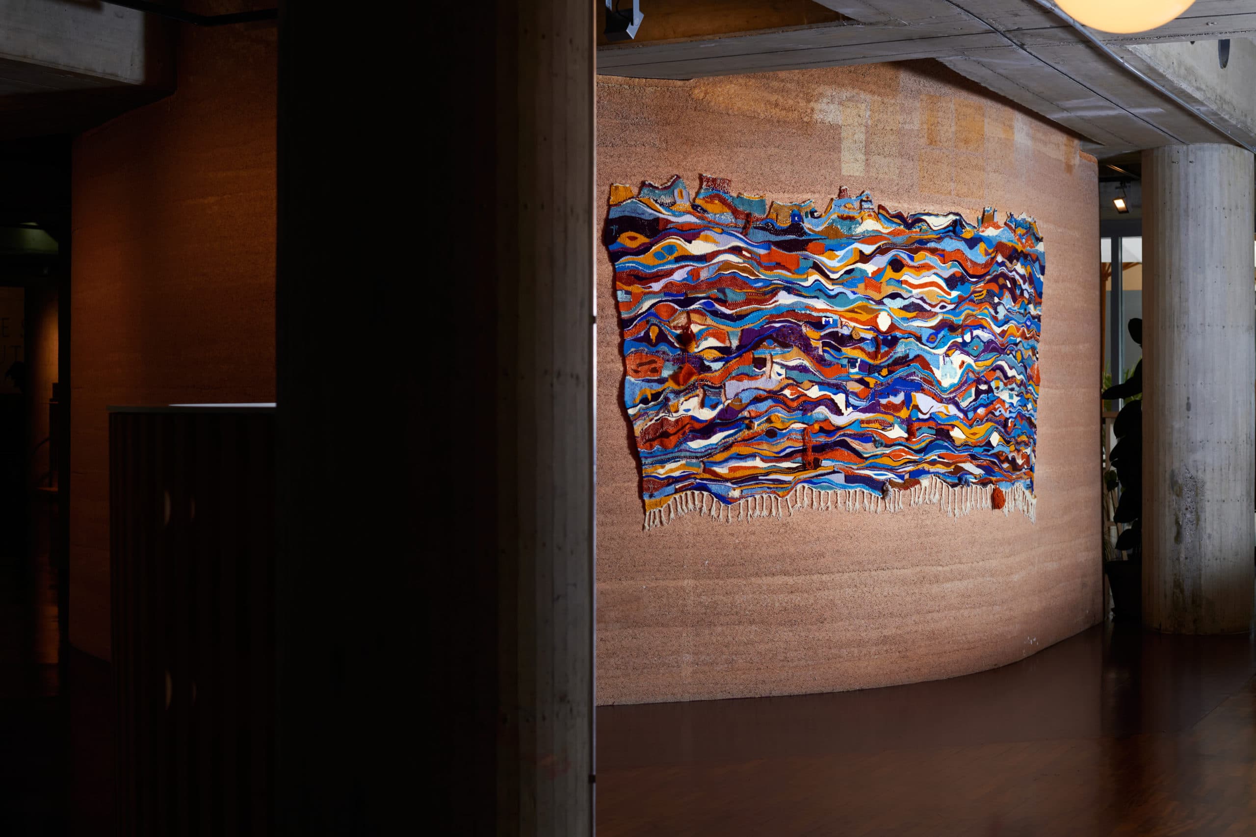 A colourful wall tapestry with abstract, undulating patterns, hung on a textured hemp wall.