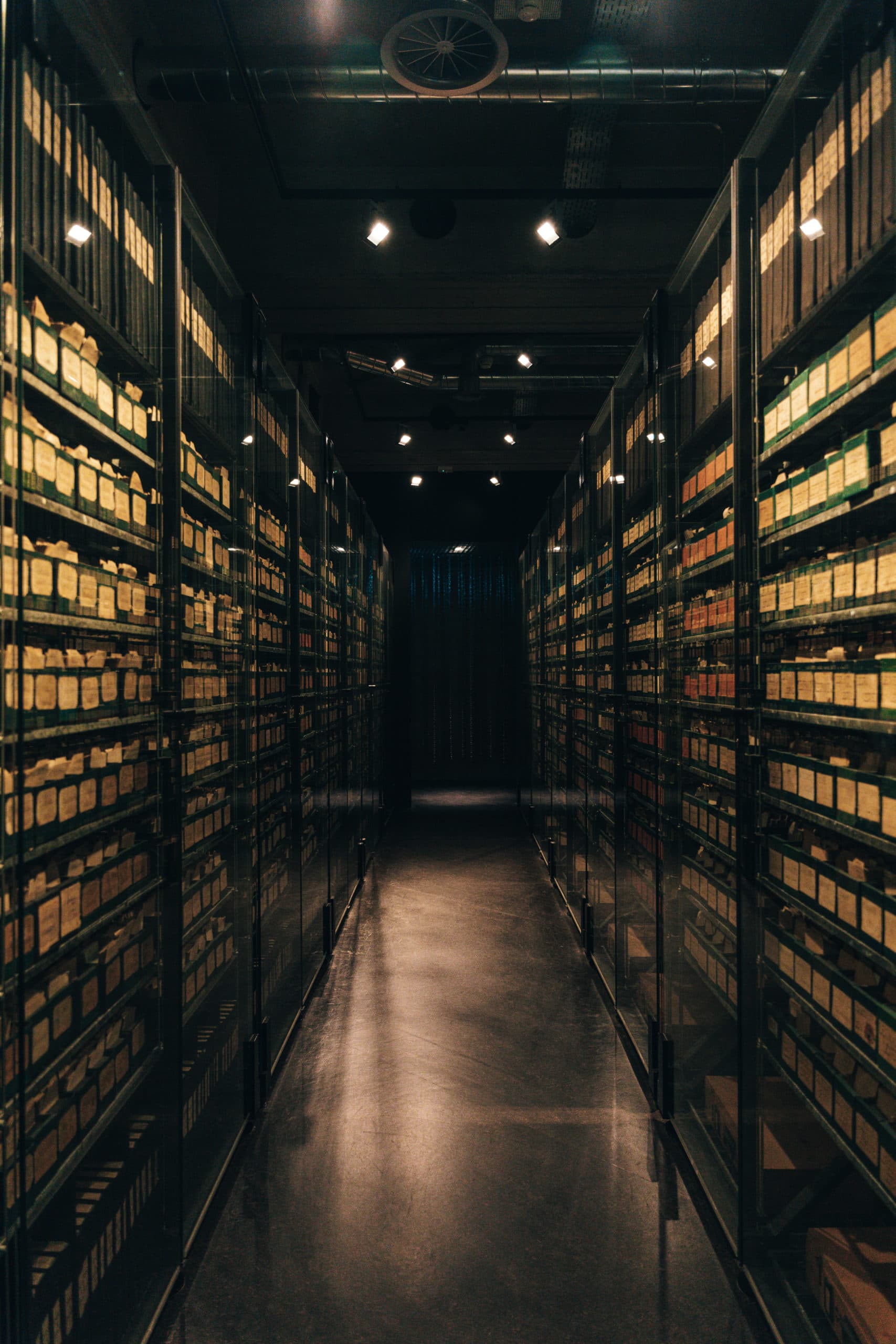 A narrow corridor lined with tall glass-fronted shelves filled with labelled archive boxes, bathed in subdued lighting.