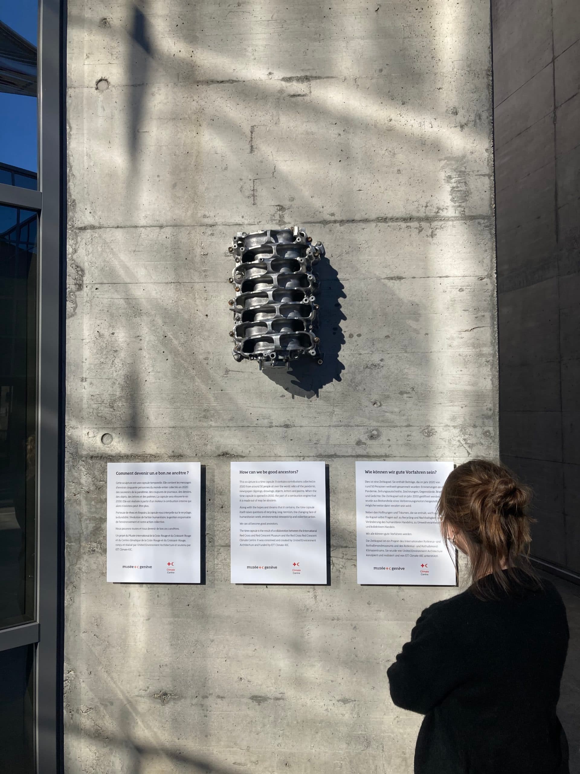 A visitor reading exhibition panels beneath a metal sculpture resembling a car engine, fixed to a concrete wall.