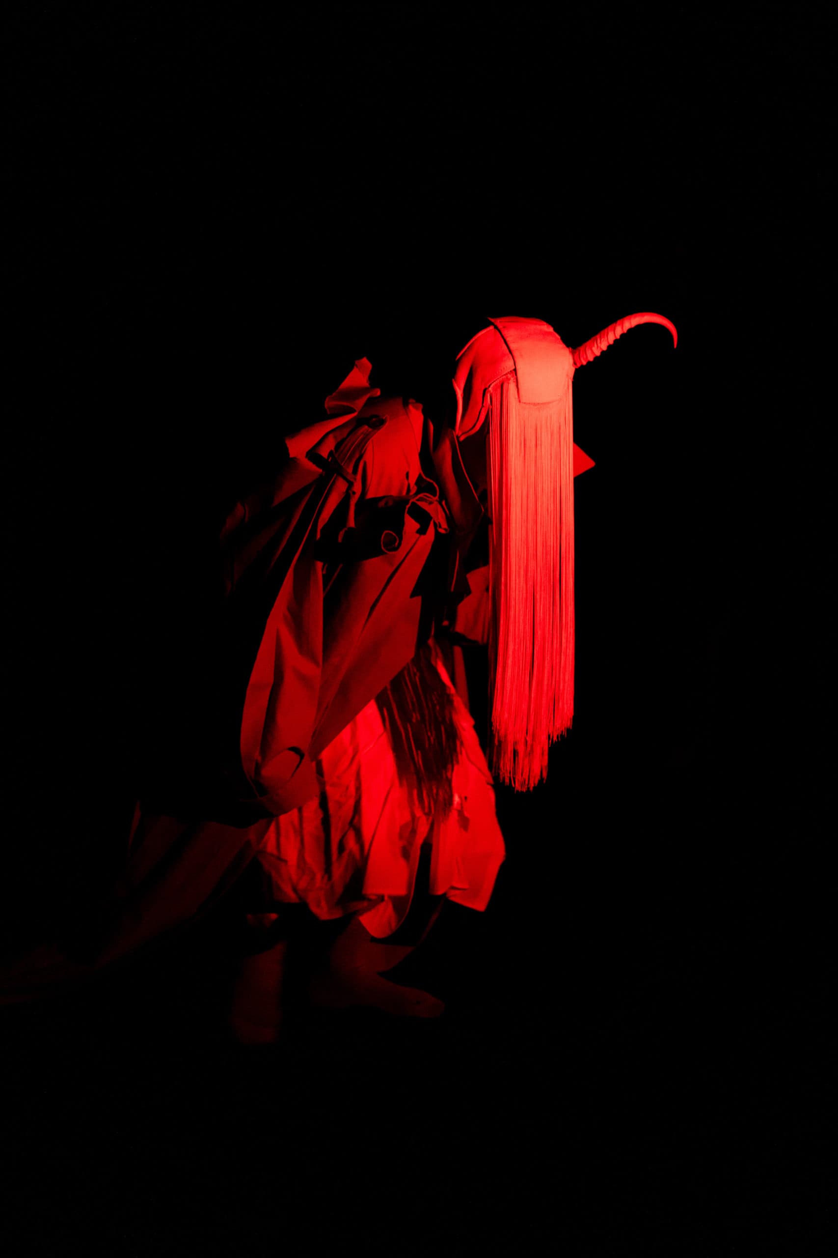 Silhouette d’une personne en costume rouge, coiffée d’un masque à corne et à longues franges, éclairée par un projecteur rouge vif sur fond noir.