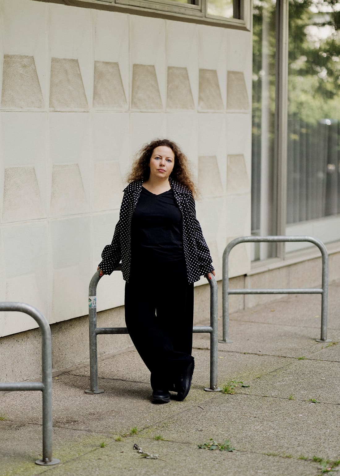 Une femme aux cheveux longs et ondulés habillée en noir est debout appuyée sur des arceaux métallique gris.