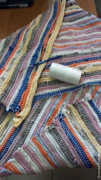 Striped multicolored woven rug in progress, with a spool of white thread and a crochet hook placed on top.