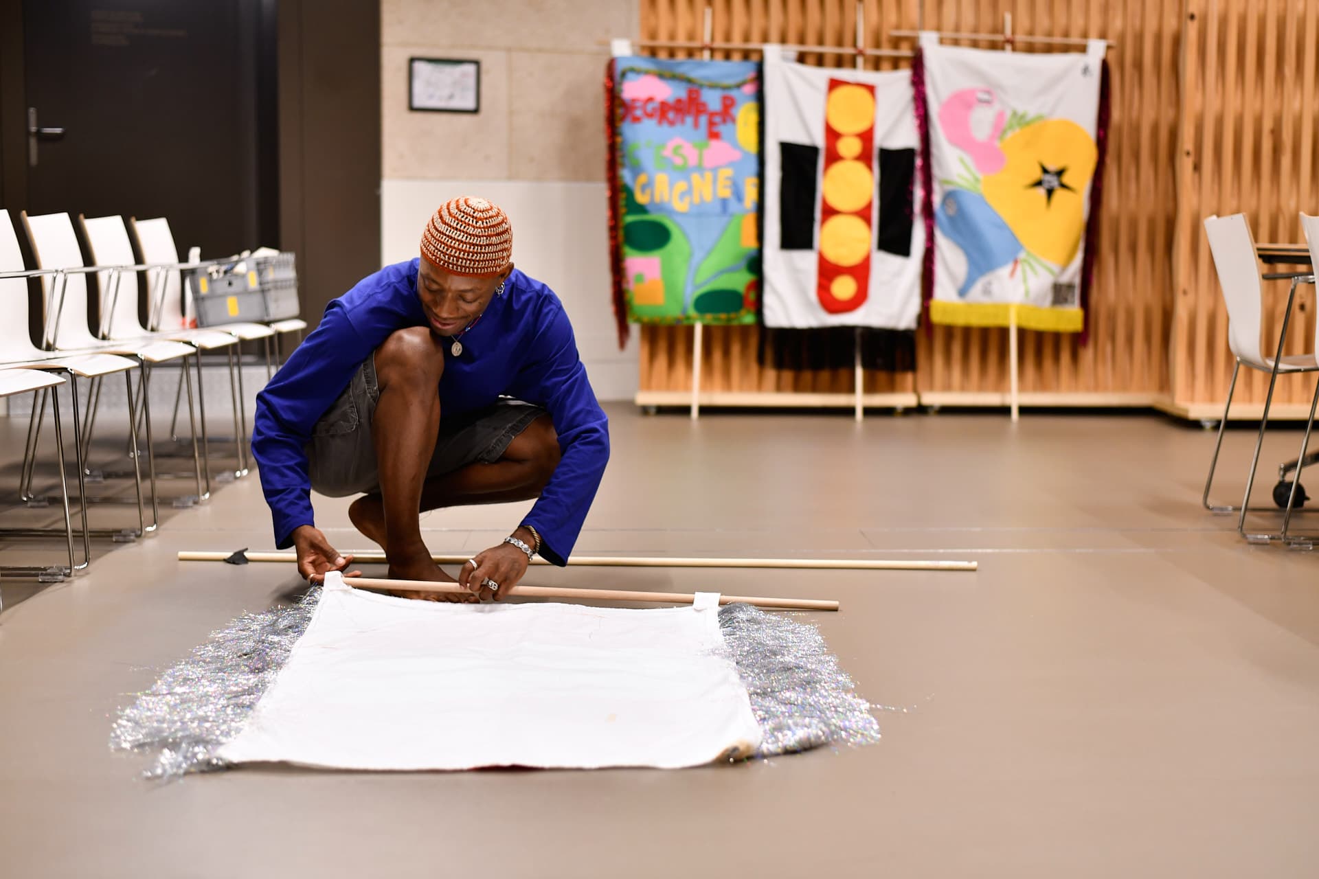 A man crouching in a room is attaching a banner to a wooden frame. Behind him, three other banners on flagpoles are leaning against a wooden wall.