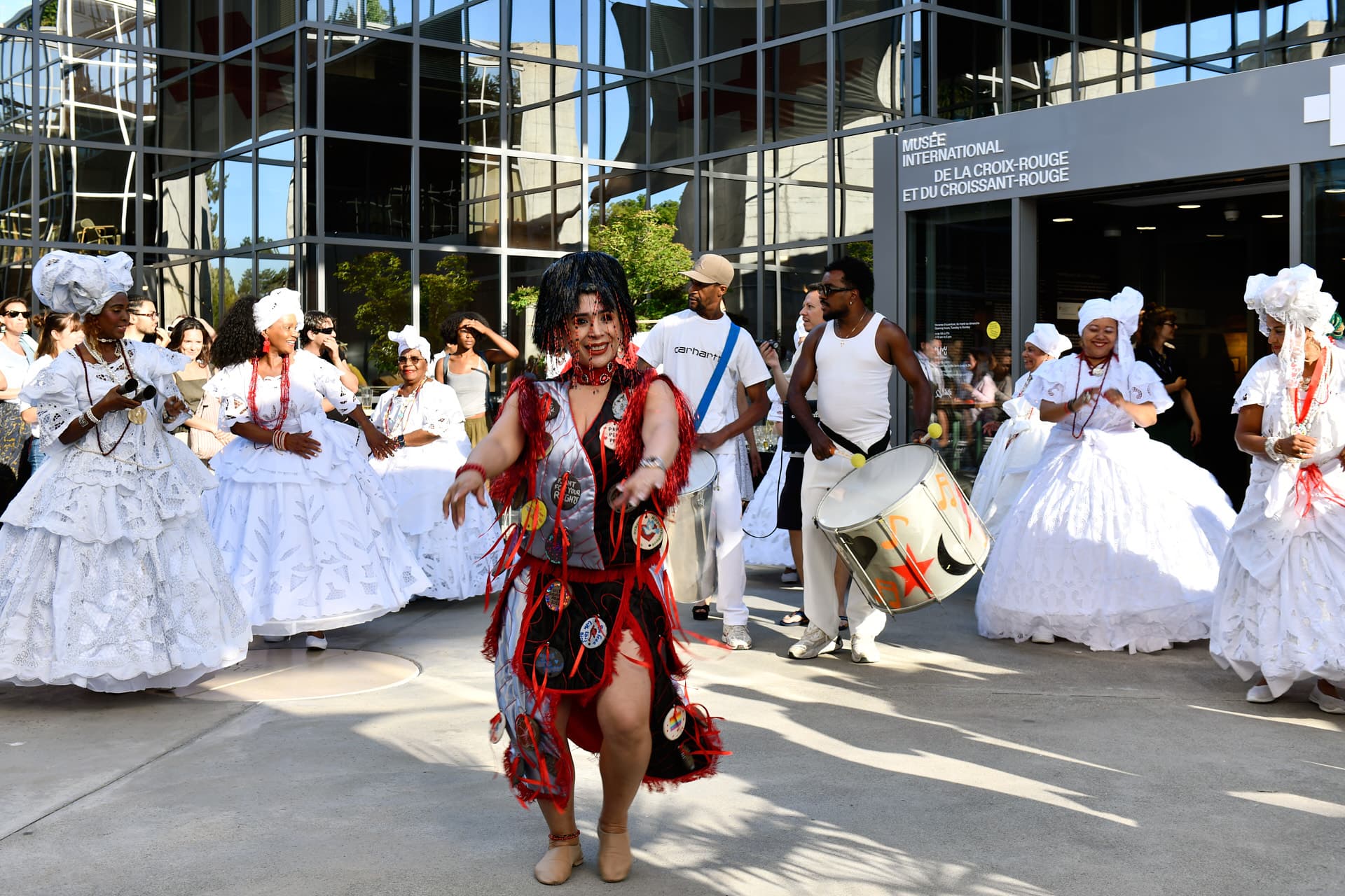 A dancer in a red and black costume is leading a festive procession, surrounded by percussionists and people wearing traditional white dresses, in front of a modern building.
