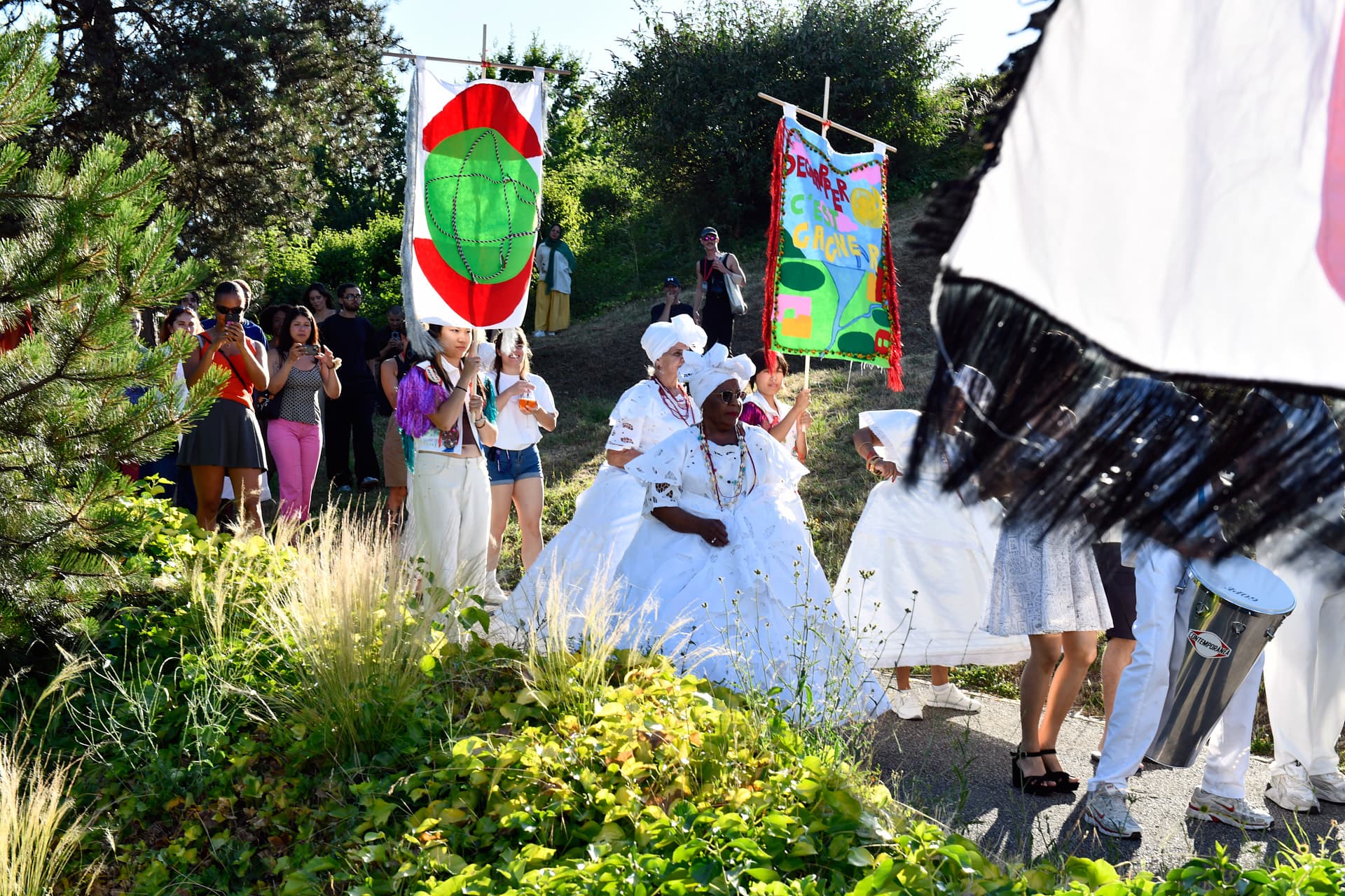 A cultural procession winds its way through a garden, with participants in traditional white robes carrying colourful banners and celebrating in the open air.