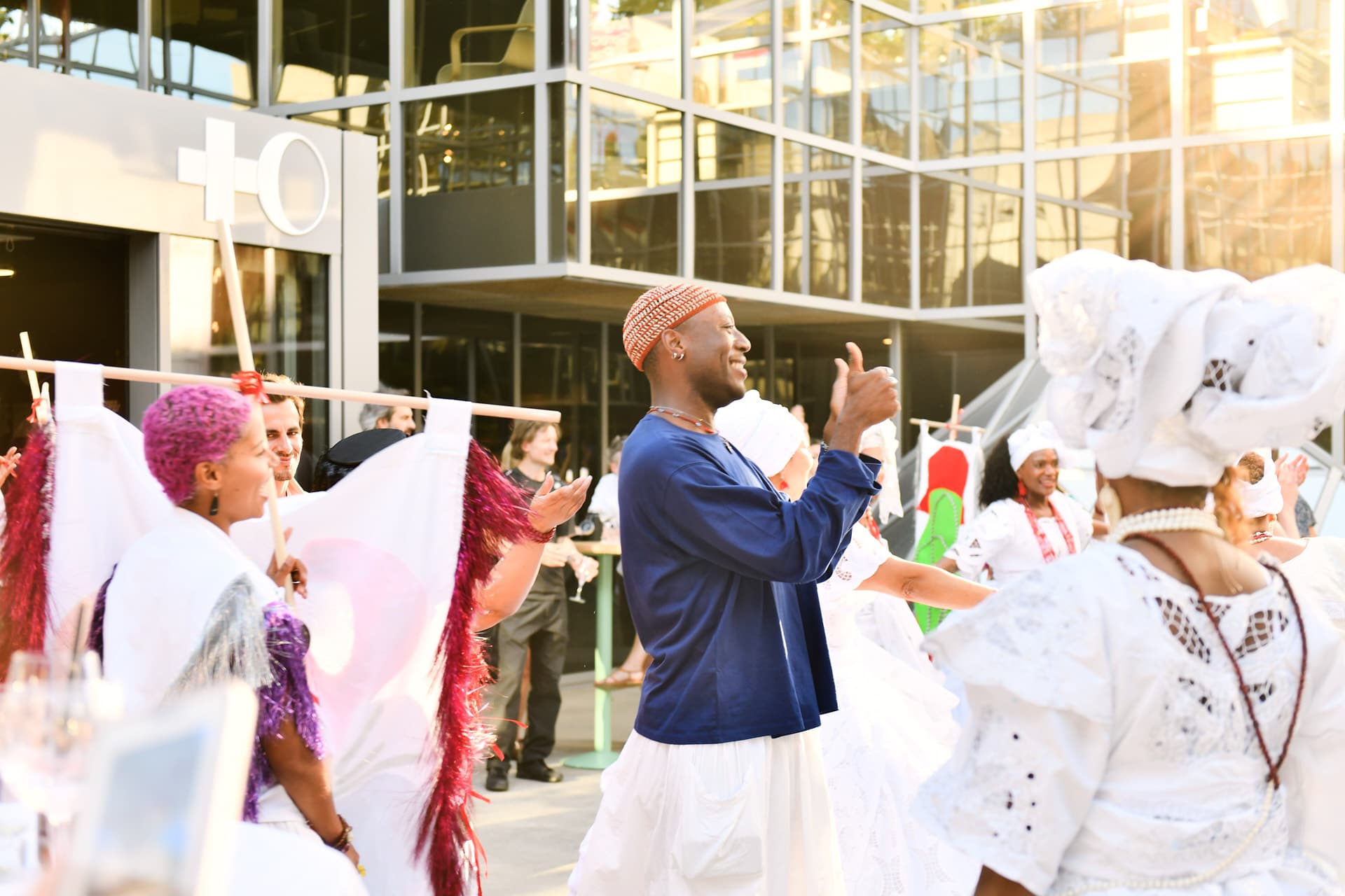 Participants in traditional white and colourful outfits are dancing and clapping in front of a glass-fronted building during an outdoor cultural celebration.
