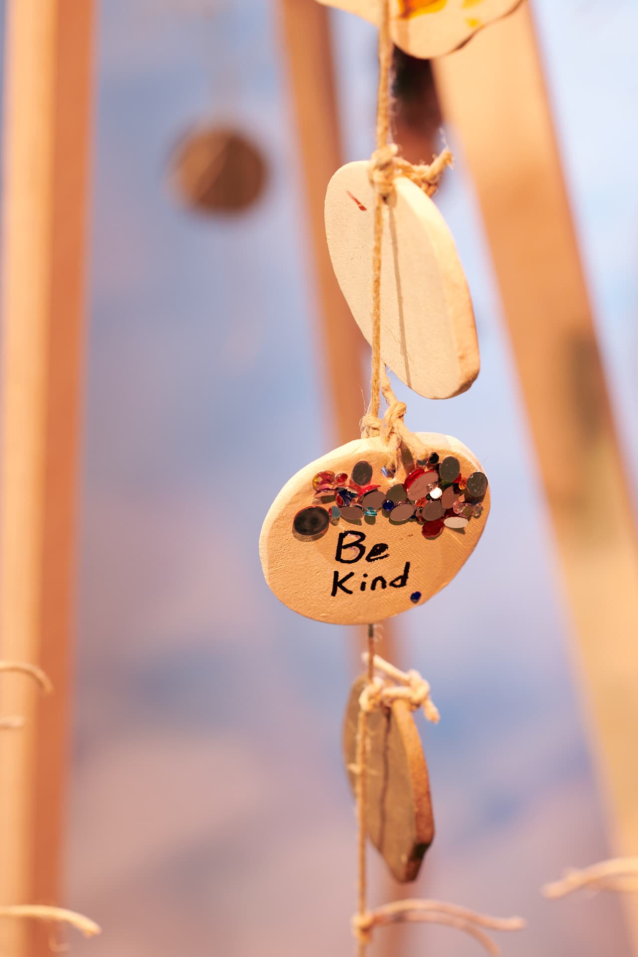 Ein runder Keramikanhänger, der zusammen mit anderen Keramikkreisen an einer Schnur hängt, mit farbigen Strasssteinen verziert ist und die Aufschrift „Be Kind“ in englischer Sprache trägt.