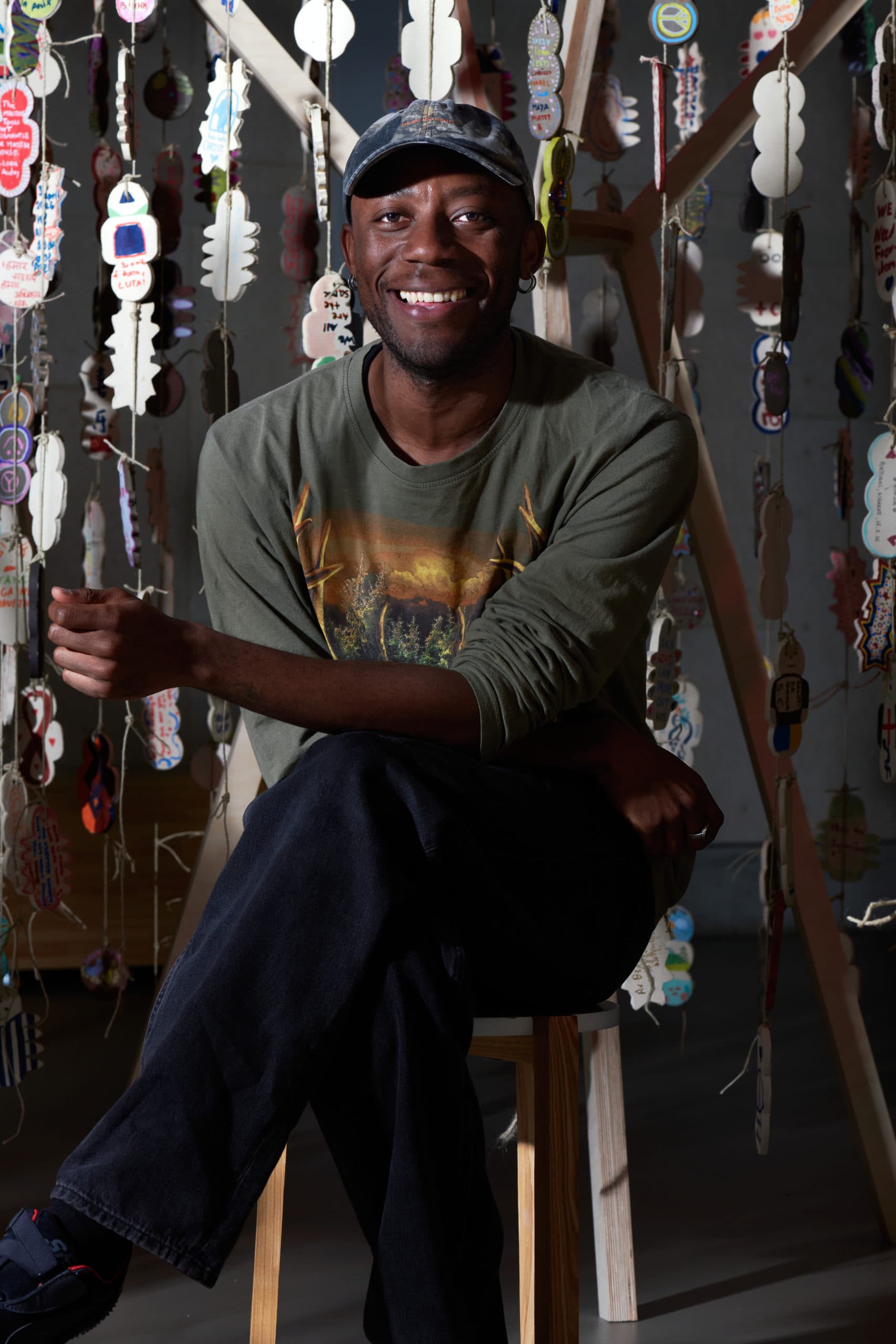 Un homme souriant, assis sur un tabouret au cœur d'une installation artistique, entouré d'amulettes en céramique suspendues, l'air détendu.
