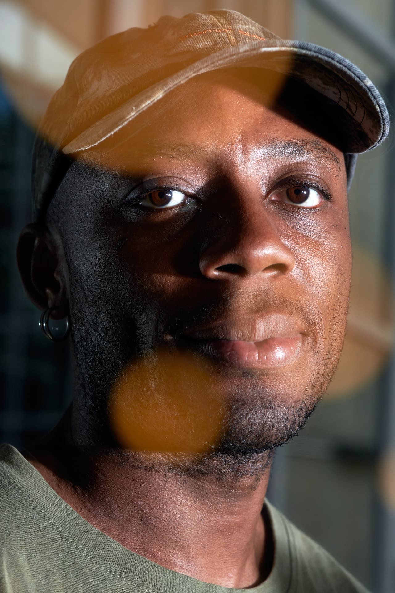 A frontal portrait of a young man wearing a cap, with light illuminating the right side of his face.