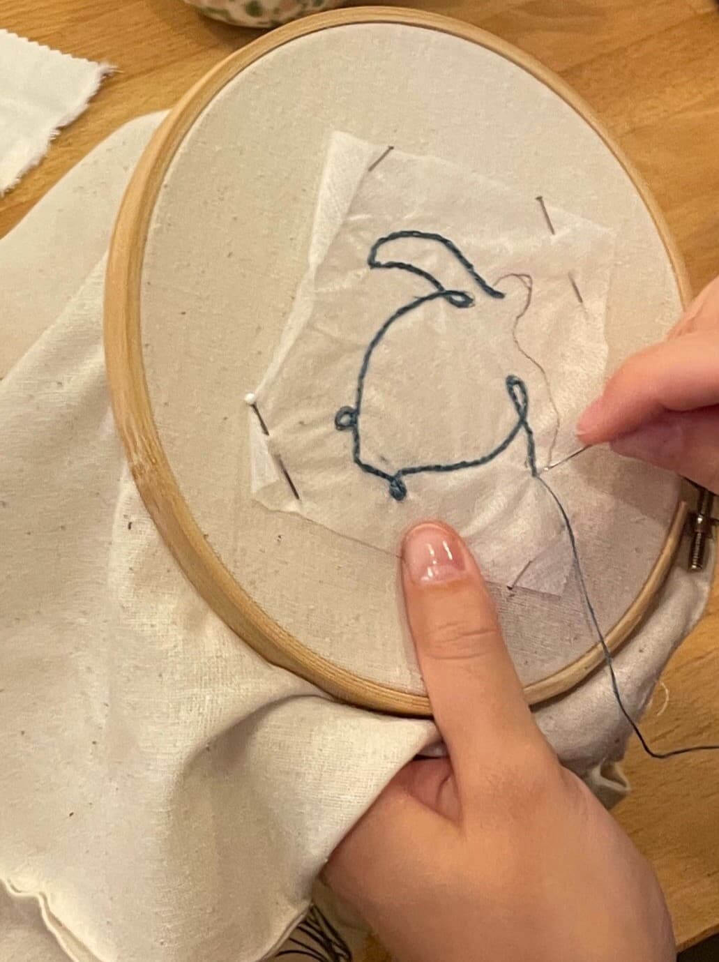 A close-up of a person’s hands embroidering the outline of a sea turtle in an embroidery hoop using navy blue thread.