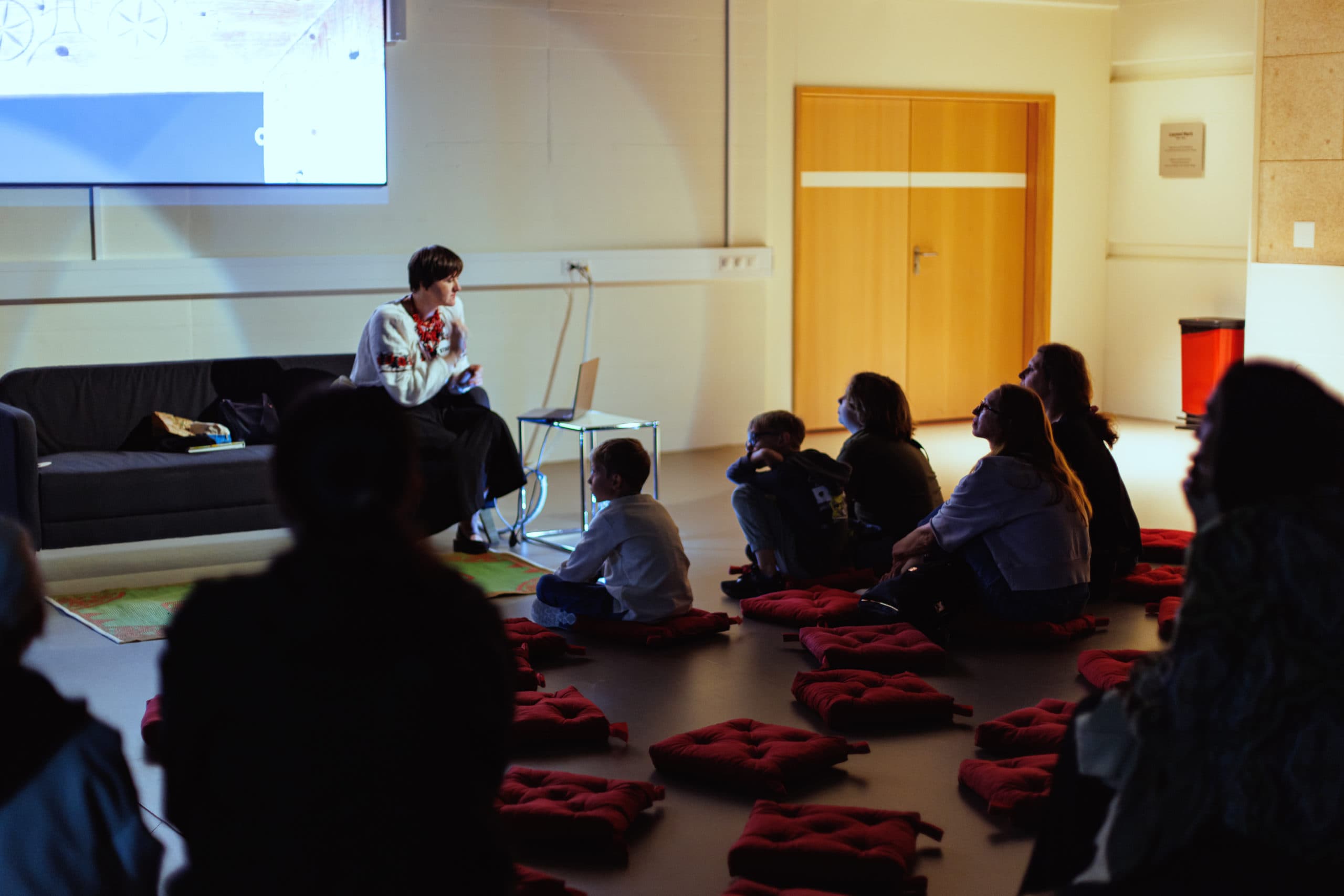 Un groupe de personnes assises au sol dans une salle, écoutant un intervenant devant un écran de projection.