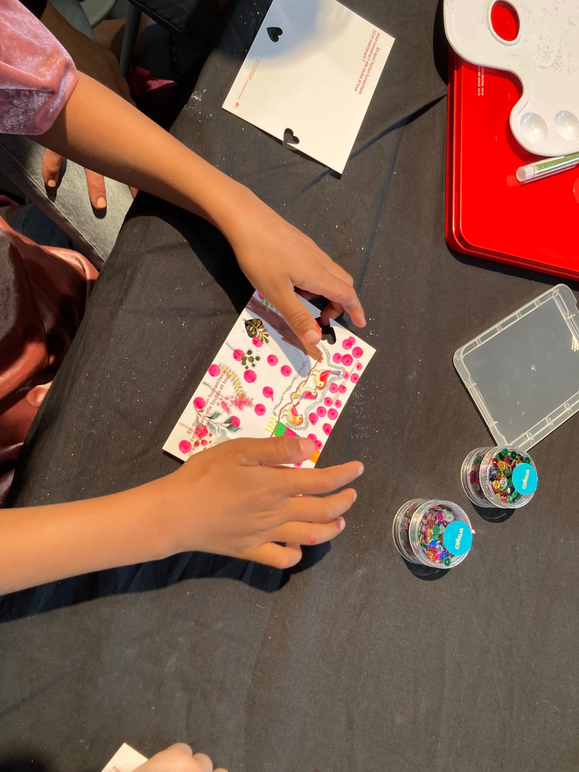 Aufnahme der Hände einer Person, die auf einem dunklen Tisch eine Postkarte mit Aufklebern und Filzstiften verziert.