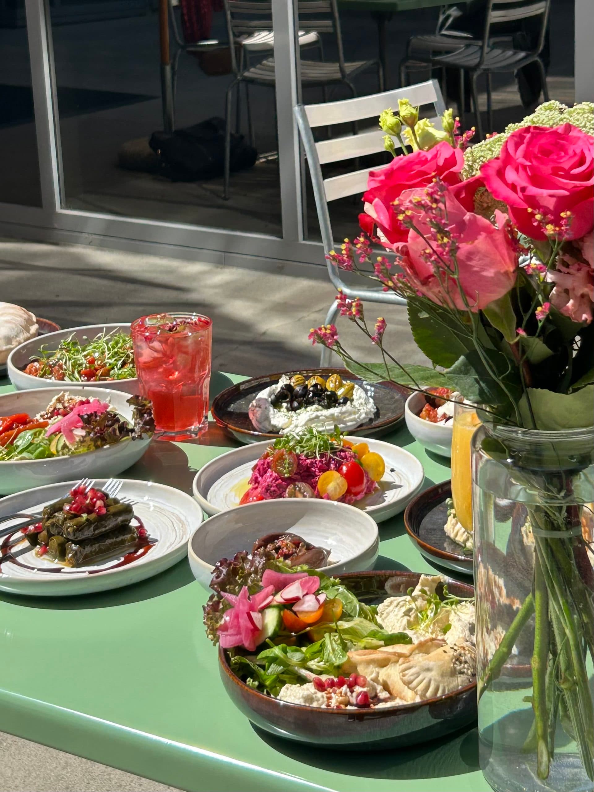 Une table verte au soleil, avec de nombreux plats colorés posés dessus et un bouquet de roses roses.