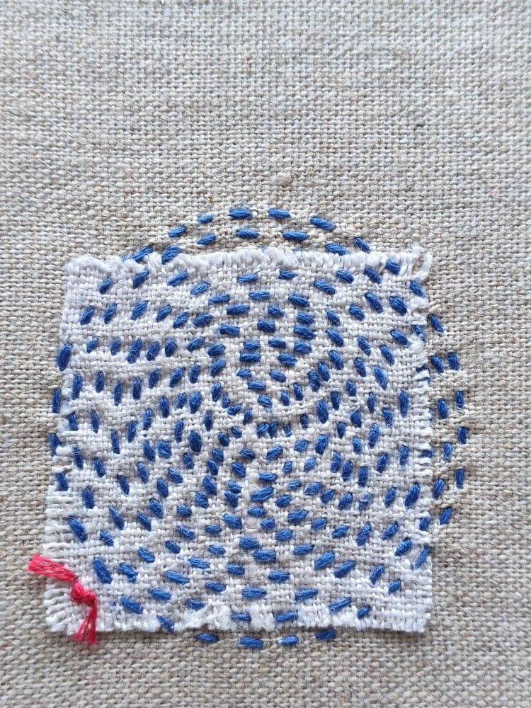 Embroidered stitches forming a circle of blue thread on grey patchwork fabric with a square of white fabric.