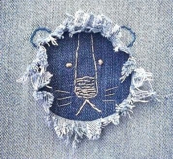 Close-up of a frayed hole in a pair of light-coloured jeans, revealing dark blue fabric with white decorative embroidery forming an animal’s face within the hole.