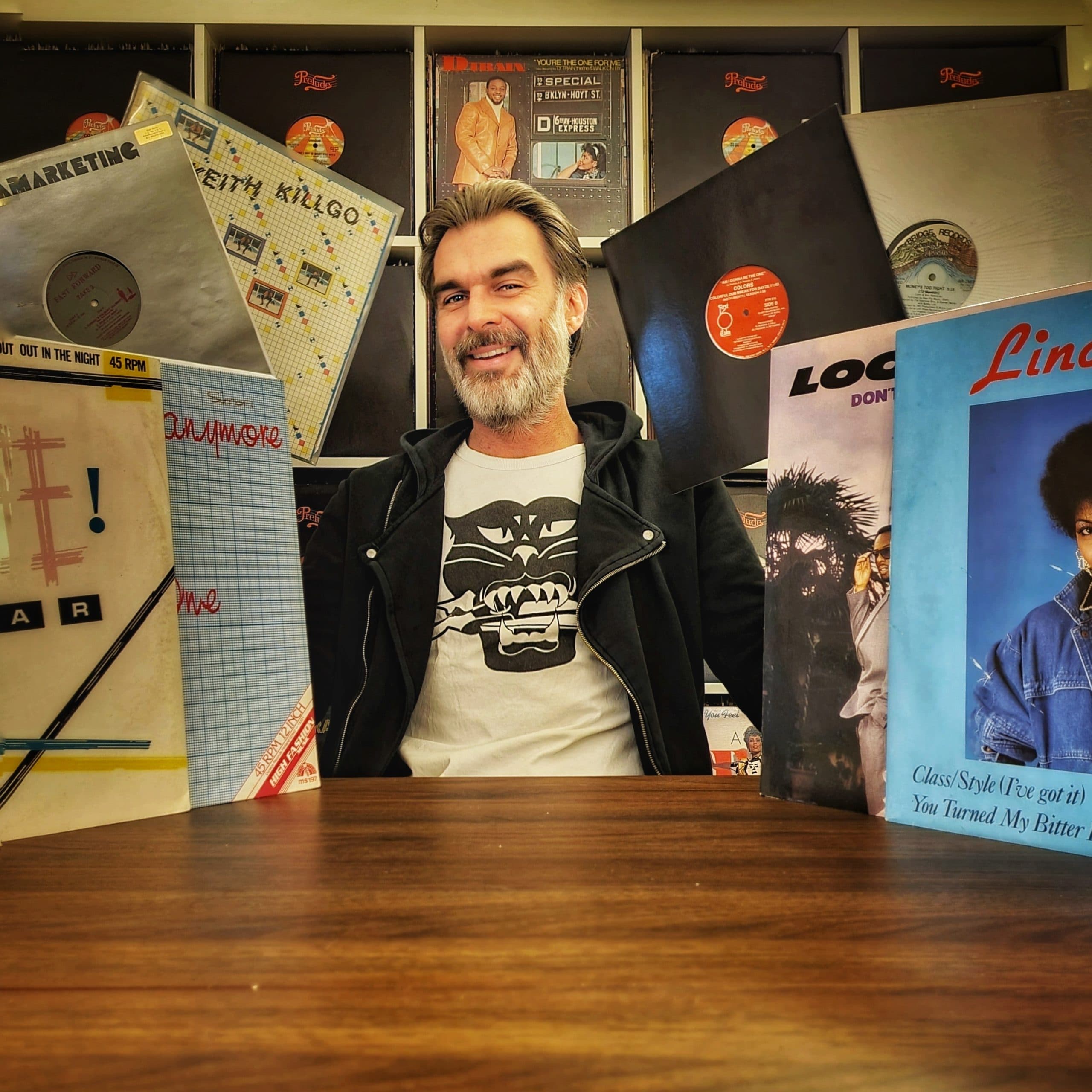 Un homme assis à une table en bois brun entouré de pochettes de vinyles ainsi que sur le mur derrière lui.