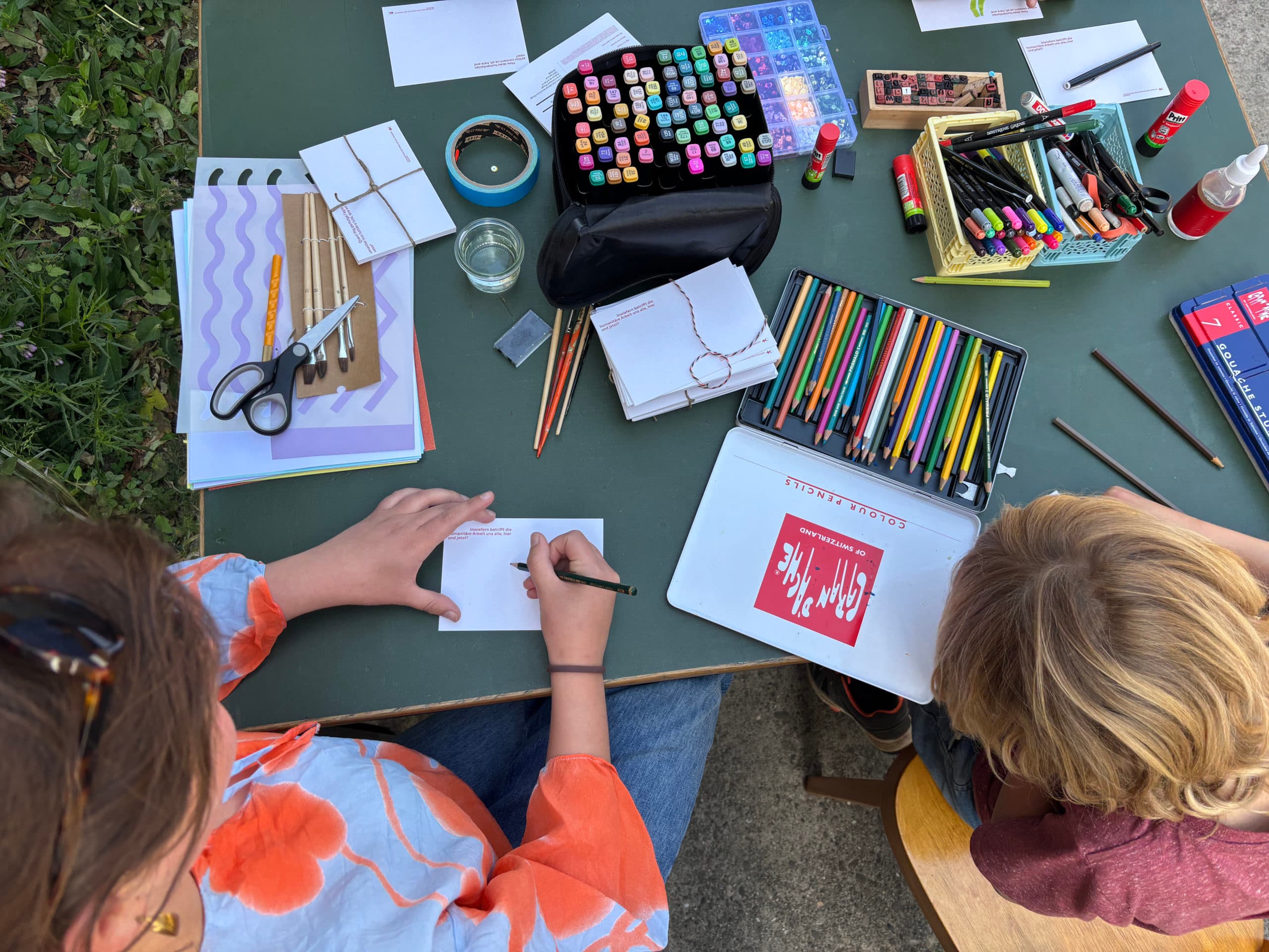 An adult and a child, seen from above, are decorating blank postcards on a table covered with drawing and craft materials such as pencils, glue and felt-tip pens.