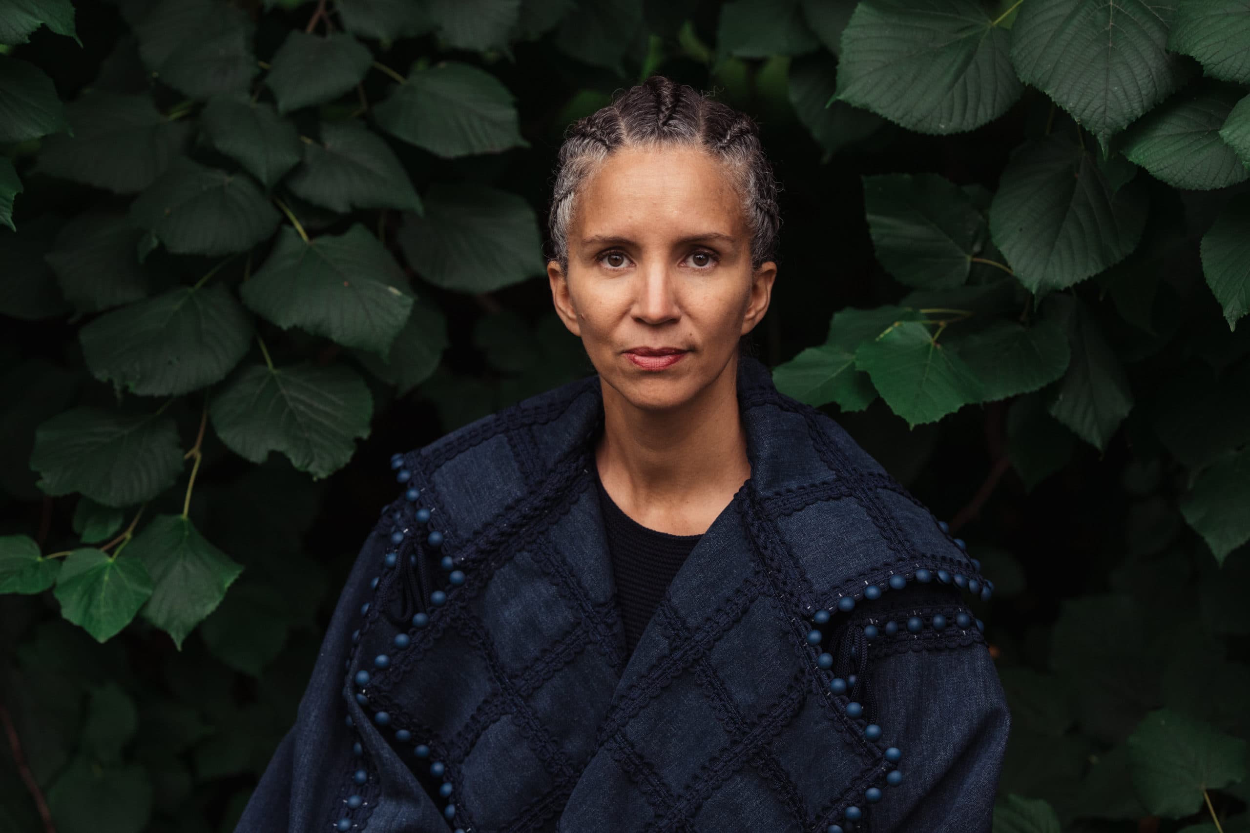 Portrait of a woman with braided hair and dark clothes in front of dark green vegetation.