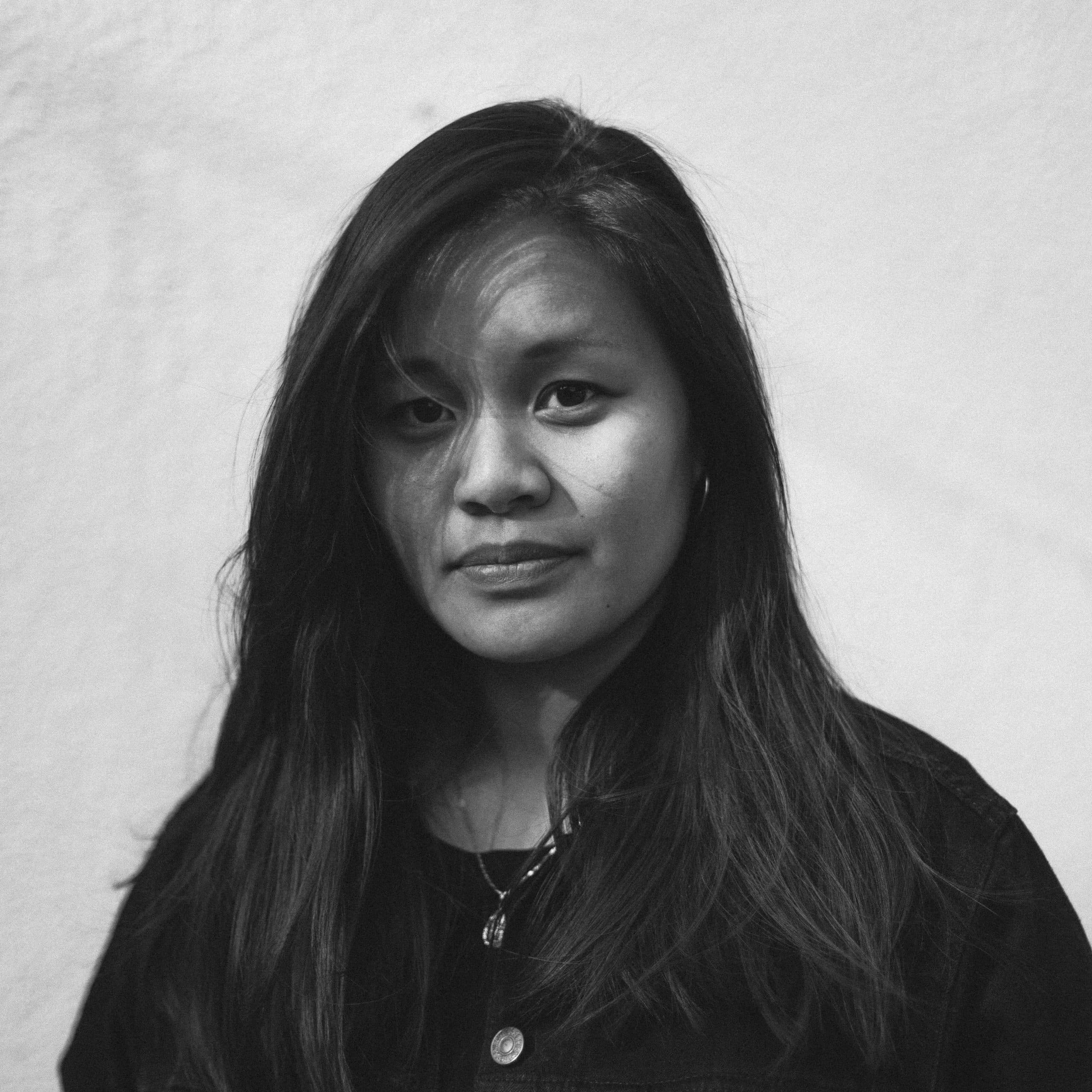 Black and white portrait of a woman with long black hair in front of a white wall.