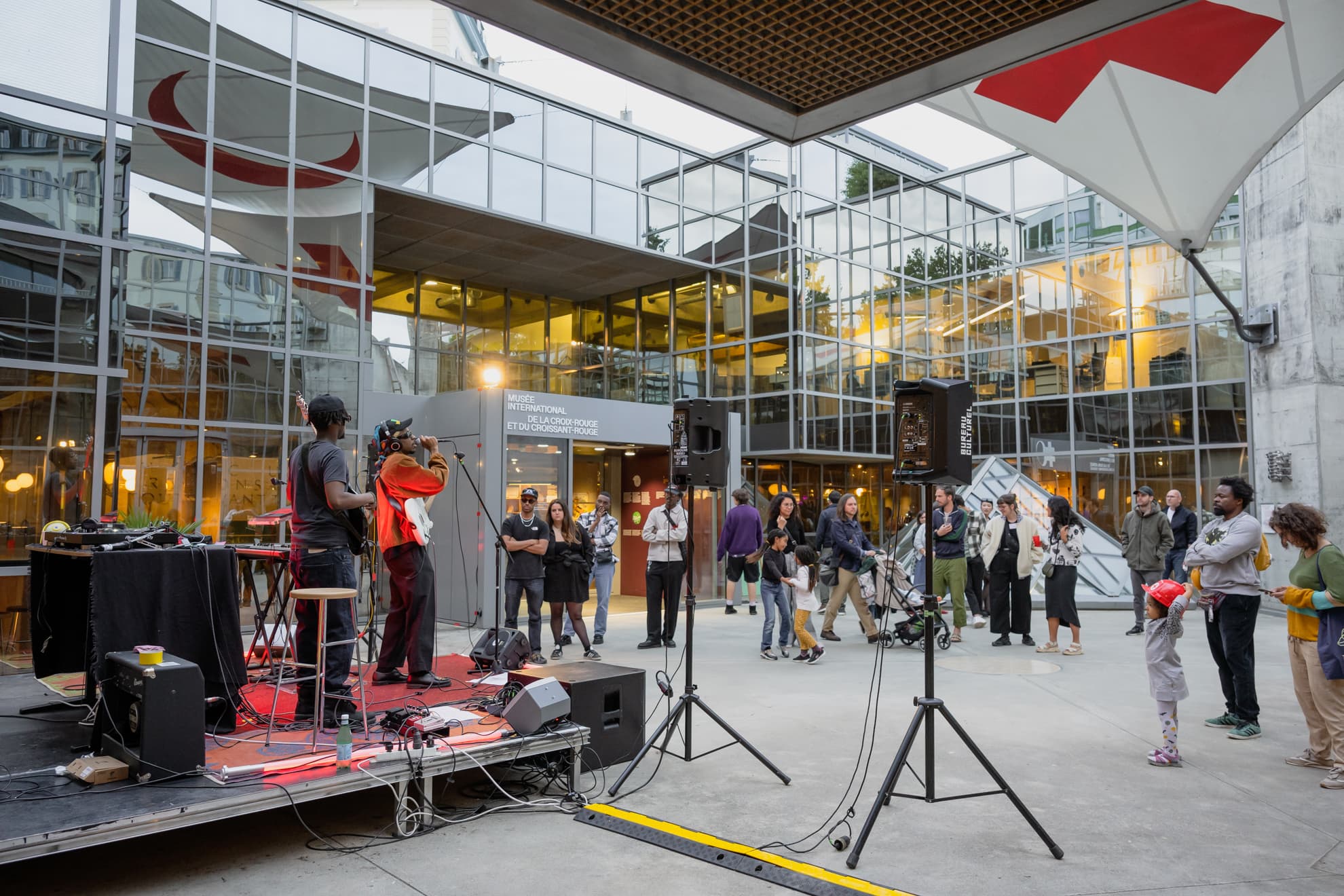 People of all ages listening to a concert in the inner courtyard of a glass-fronted building.