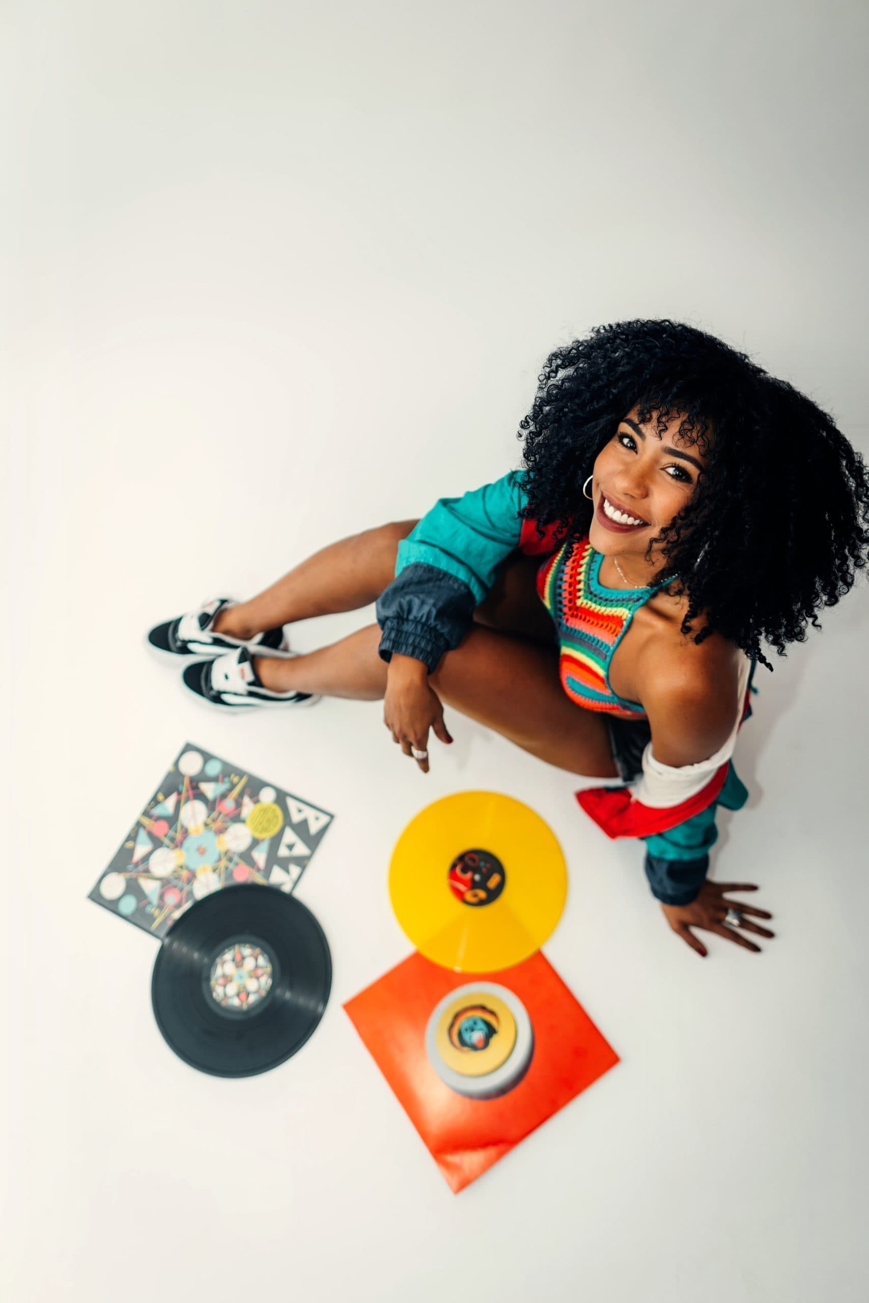 A smiling woman dressed in colourful clothes, photographed from above, sitting on a white floor surrounded by vinyl records and colourful record sleeves.