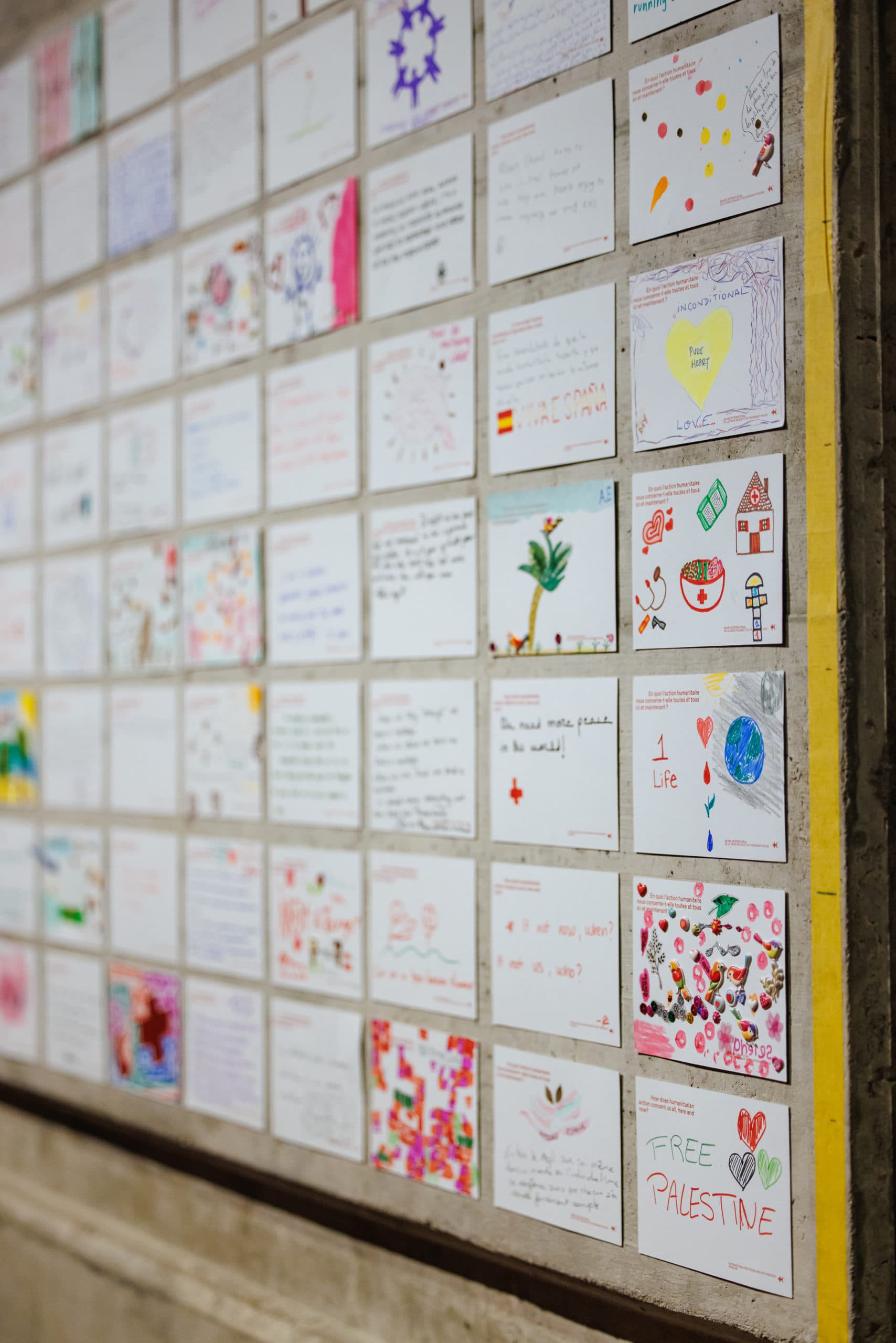 Postcards featuring drawings, colouring pictures and text in a variety of colours, stuck onto a grey wall in a regular grid pattern.