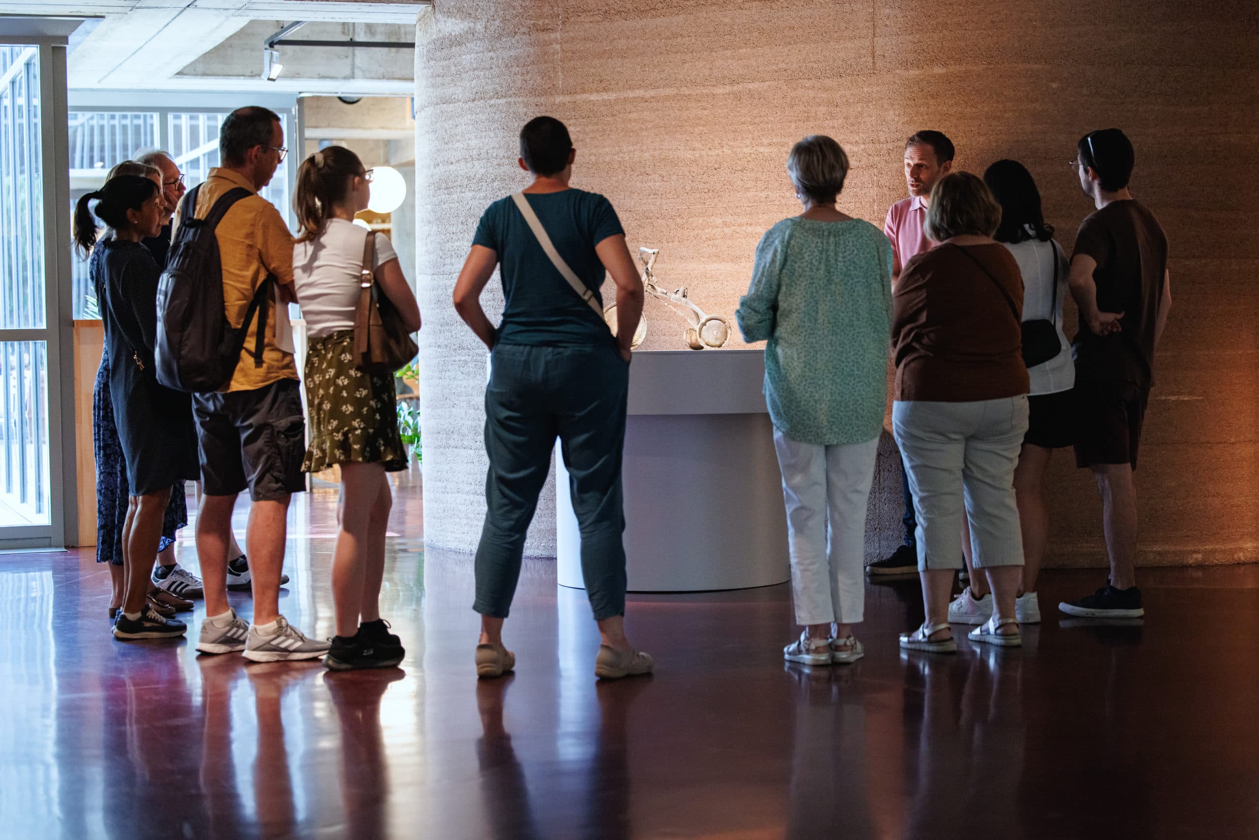 A group of about ten people are standing in an exhibition hall, gathered around an illuminated display case and listening to a presentation.